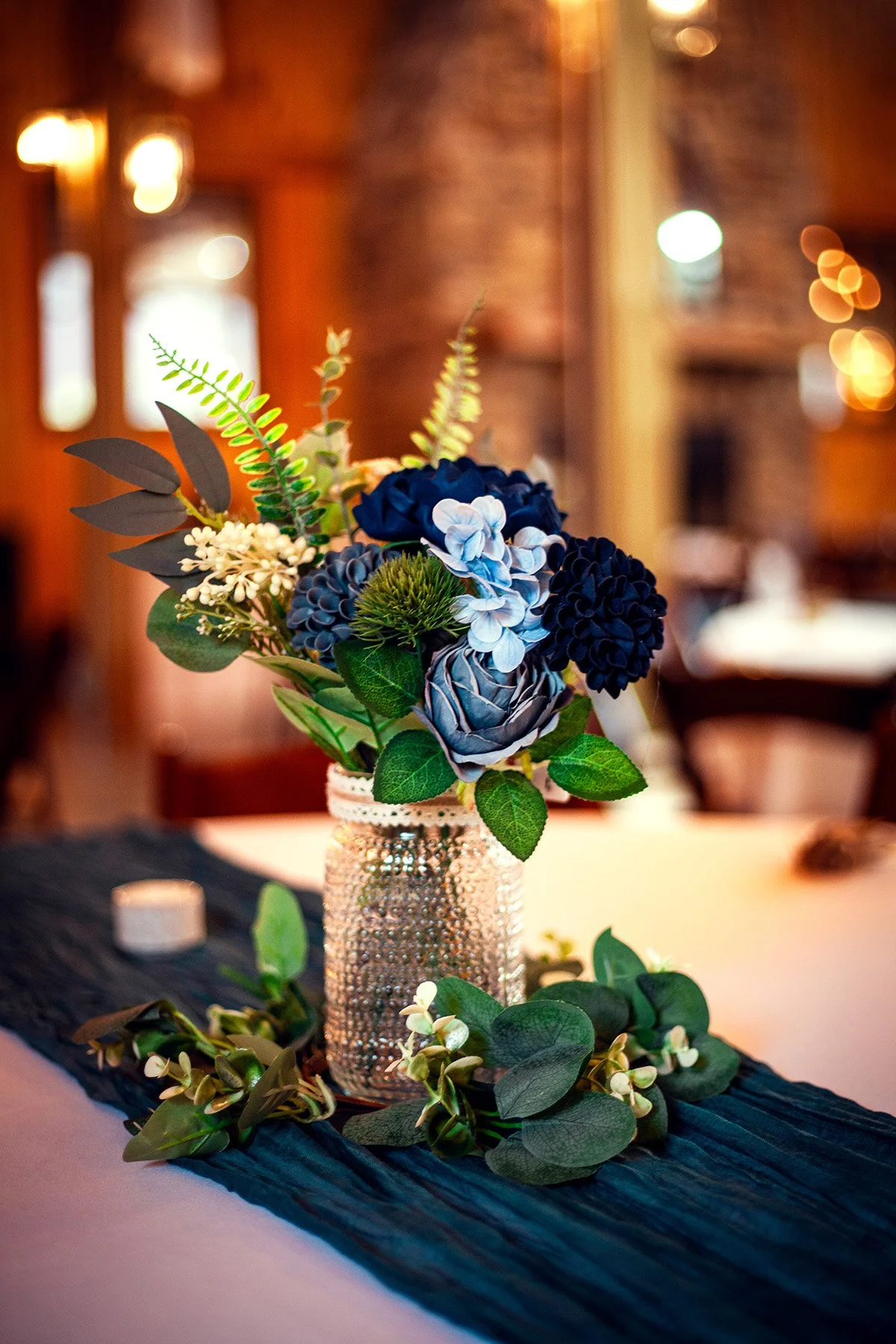A floral centerpiece with blue, white, and green flowers and green leaves in a textured glass vase on a dark table runner.