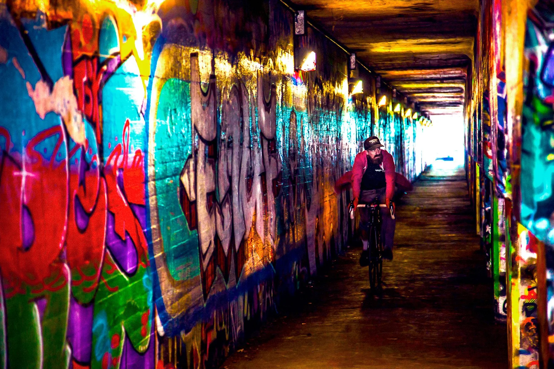A man riding a bicycle through an underground tunnel covered in colorful graffiti art, with a row of small lights along the ceiling and light at the end of the tunnel in the background.