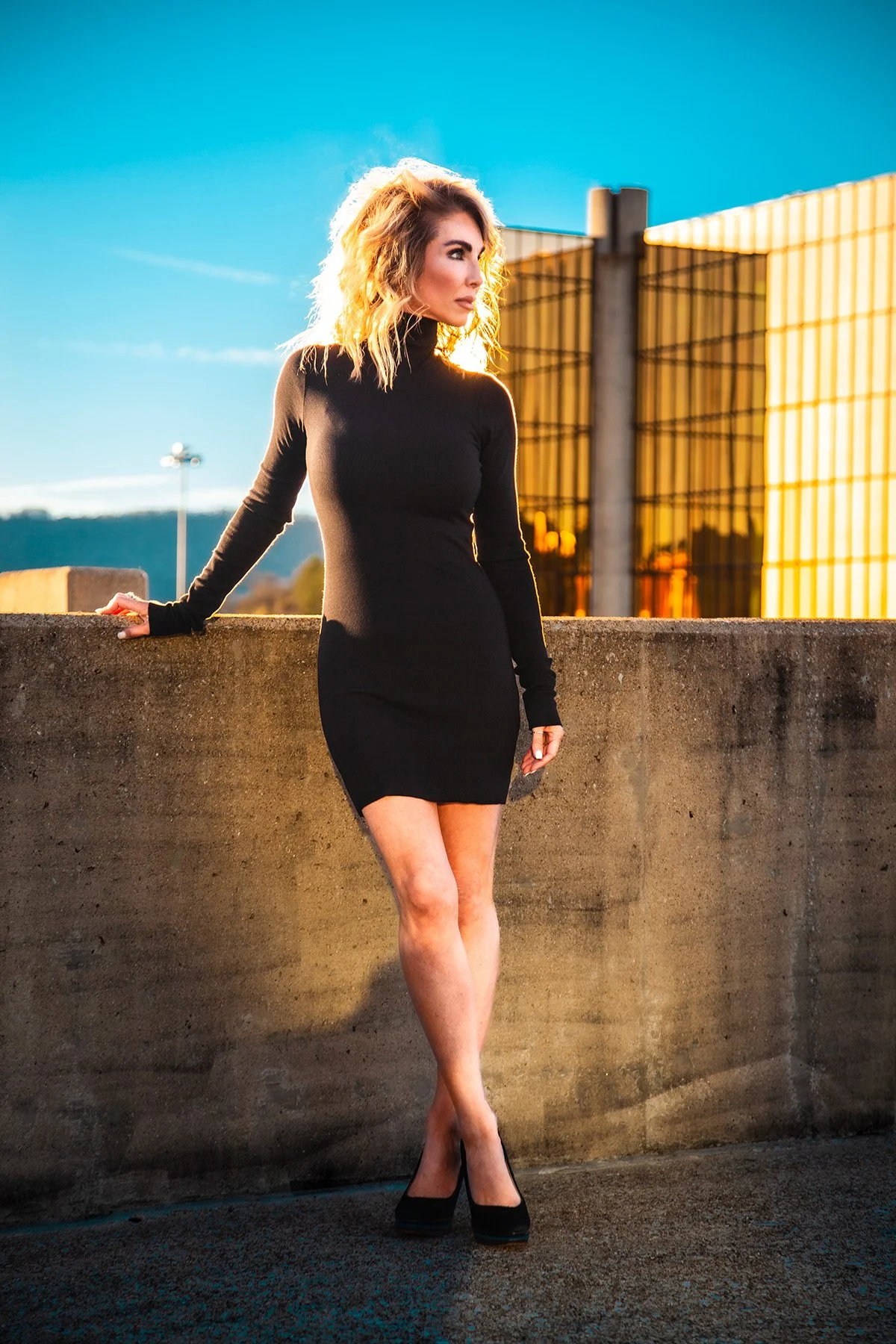 A woman in a black dress and high heels standing outdoors on a concrete ledge at sunset, with modern buildings and a hill in the background.
