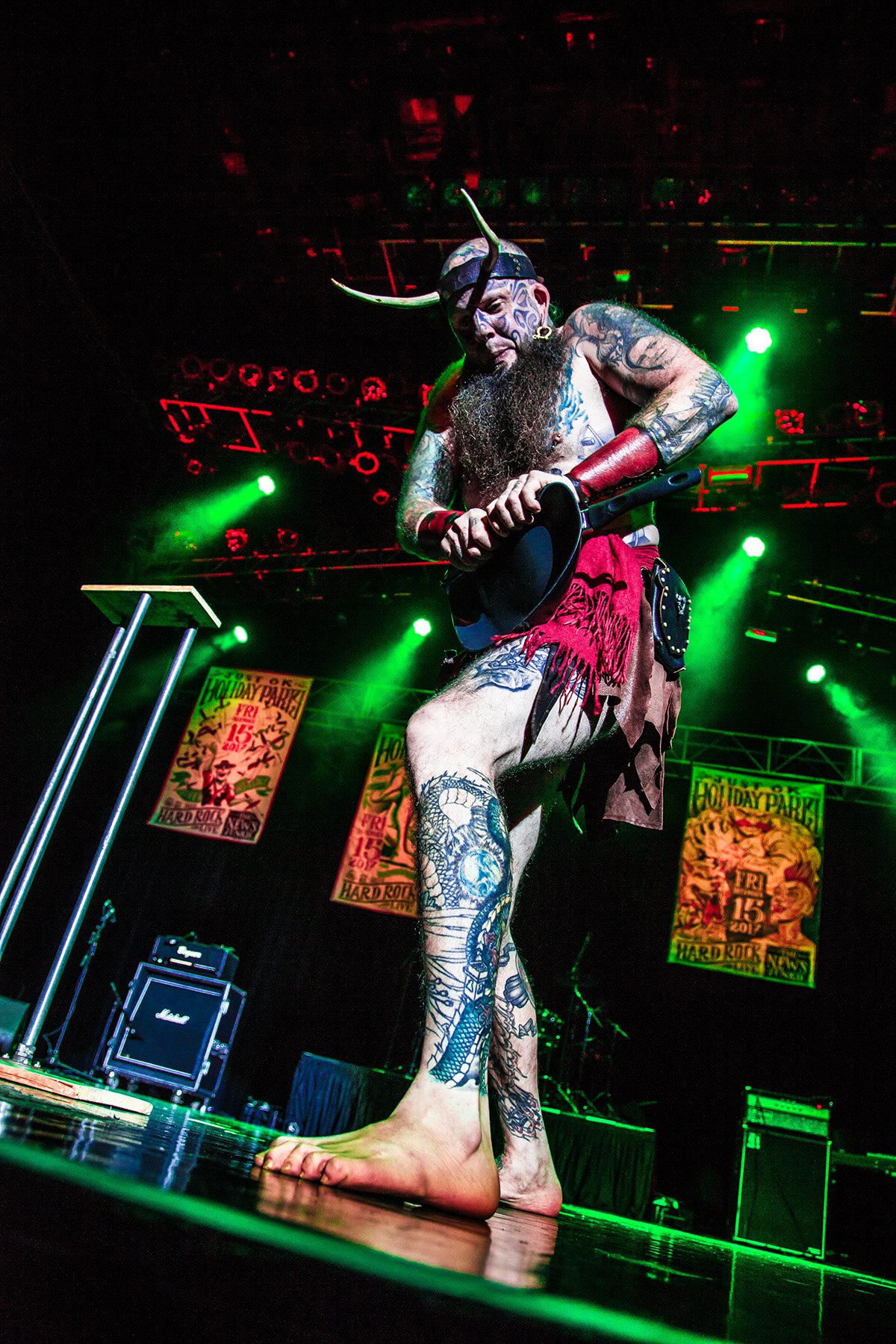 A tattooed performer with face paint, wearing a horned helmet, red arm guards, and a red loincloth, barefoot, playing the guitar on stage under green lights at a music festival.