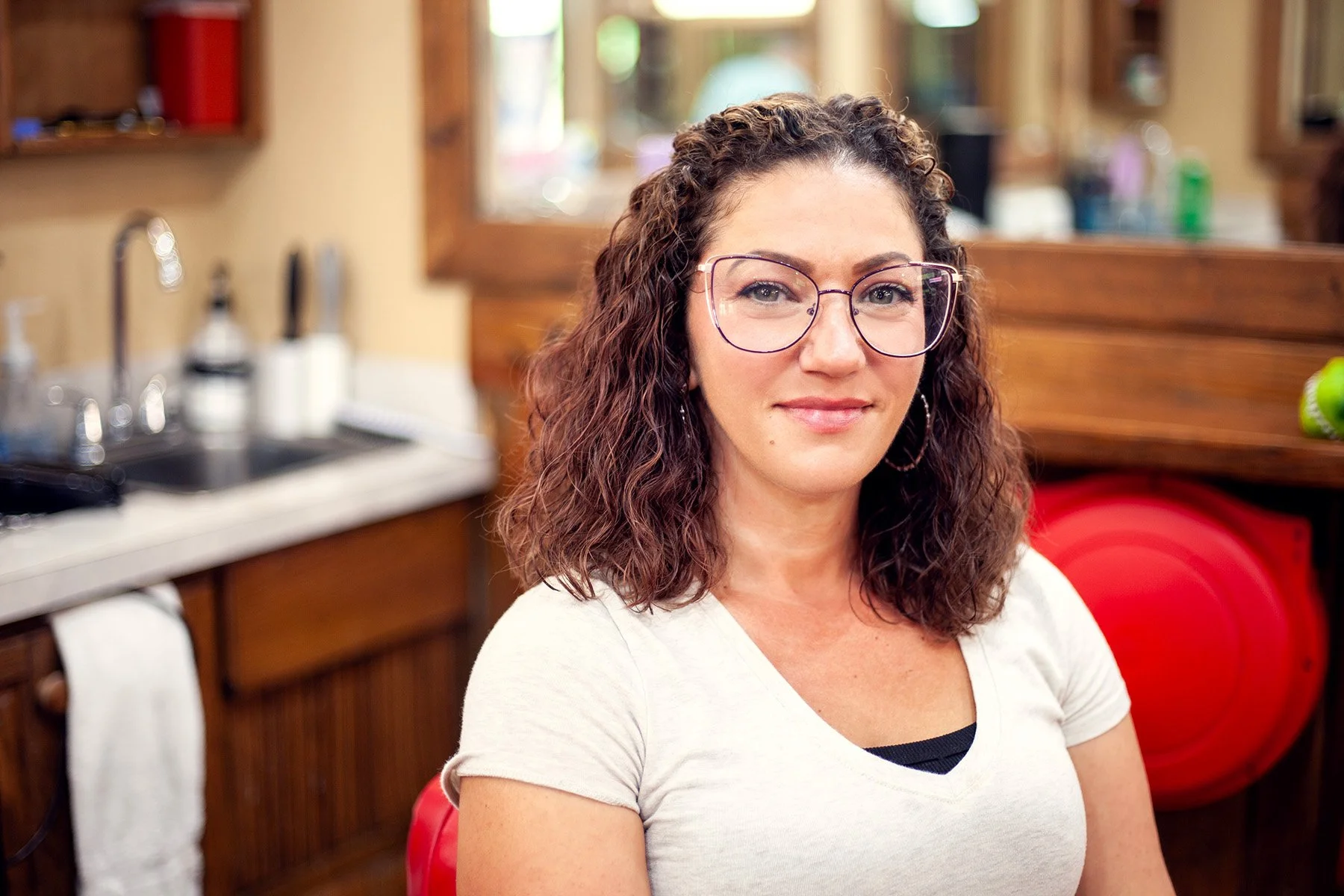 A woman with curly brown hair, glasses, and wearing a white shirt, smiling in a kitchen with wooden cabinets and a red fire hydrant in the background.