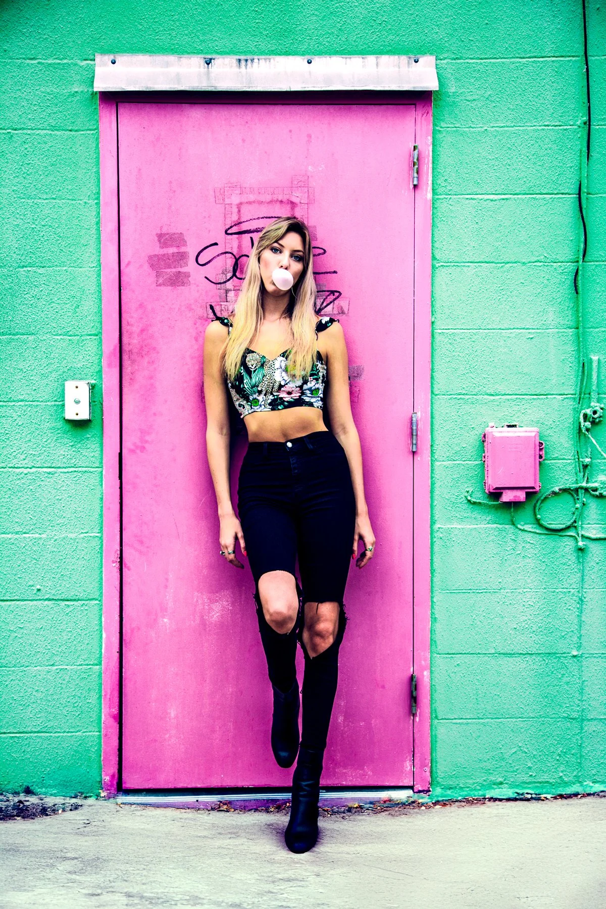 A young woman with long blonde hair wearing a floral crop top, black ripped jeans, and black boots blowing a bubble gum bubble standing in front of a pink door and green wall.