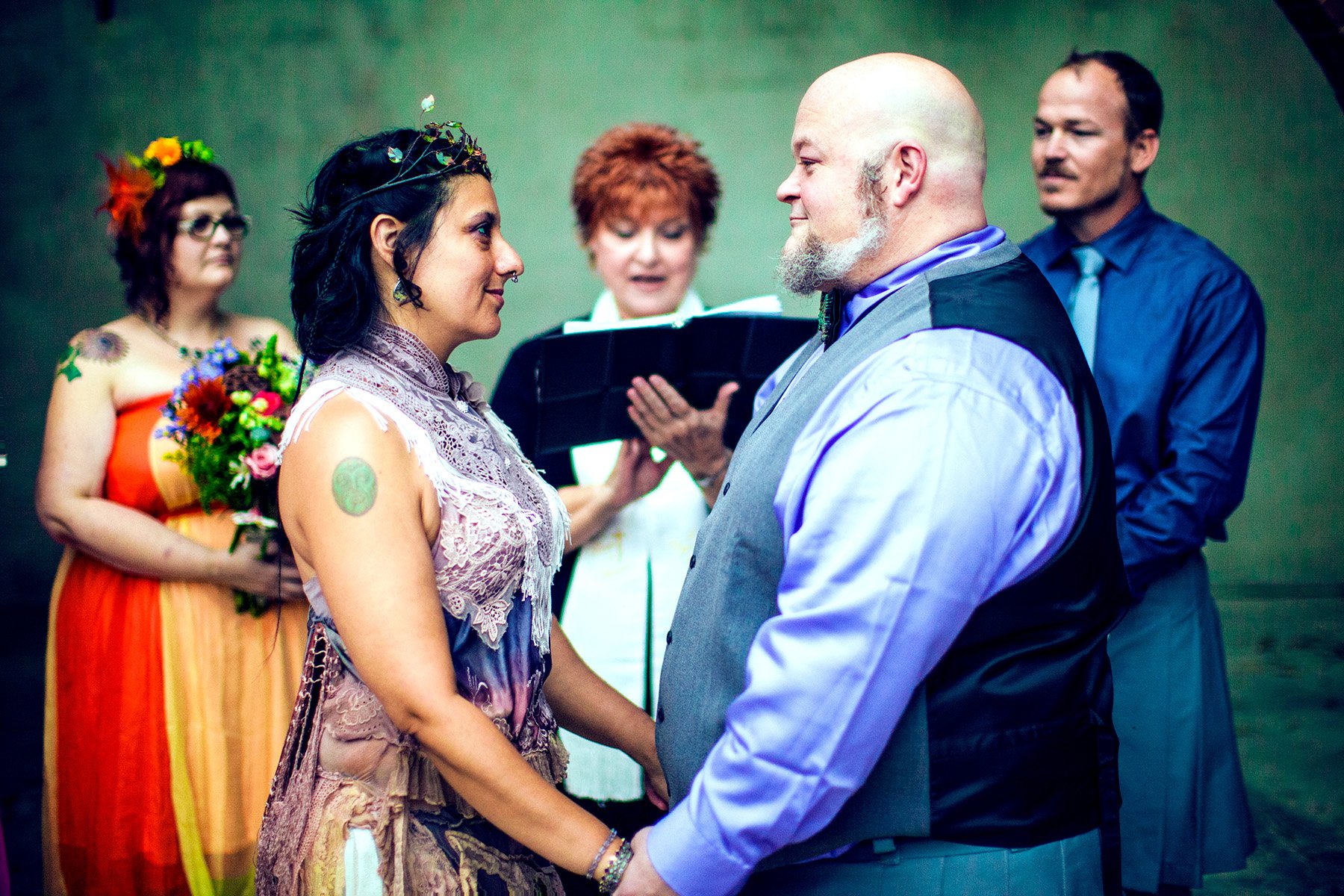 A wedding ceremony with a couple holding hands and looking at each other, surrounded by friends and officiant. The woman wears lace and has a flower tattoo on her arm, while the man has a bald head and beard. The background friends wear colorful outf