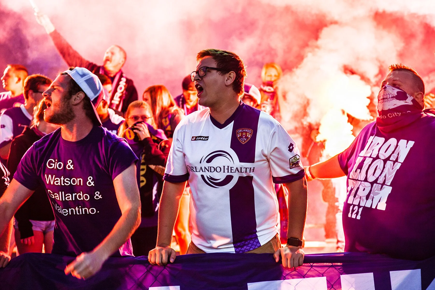 Group of Orlando City soccer fans celebrating with smoke and passionate expressions. Central figure wearing Orlando City jersey with orange, purple, and white colors.