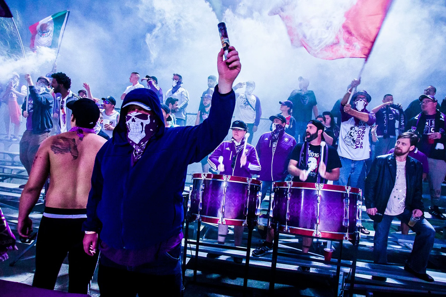 Group of sports fans in a stadium, some wearing masks, some holding flags, and others playing drums, with smoke and vibrant lighting.