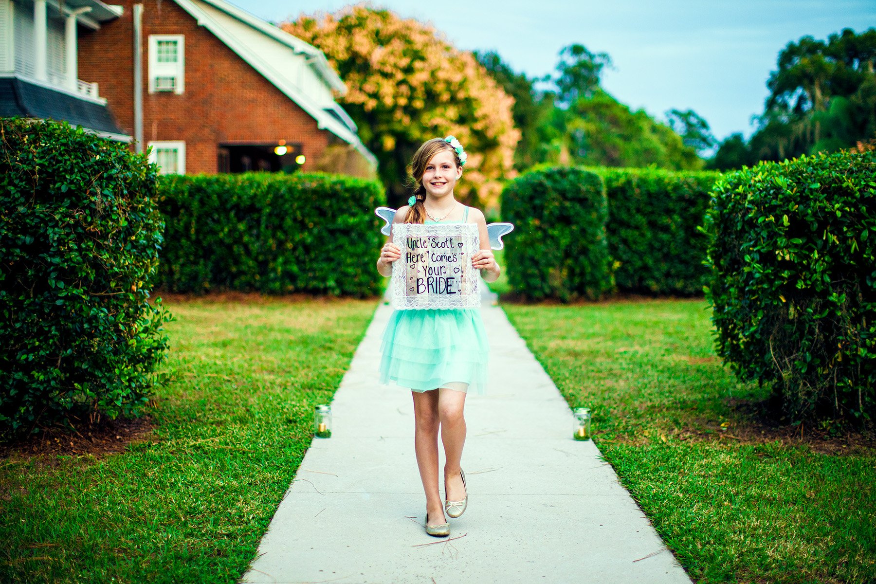 A young girl in a light green dress holding a sign that reads 'Uncle Scott, here comes your bride' walks down a sidewalk flanked by hedges leading to a house with a brick wall and white roof in the background.
