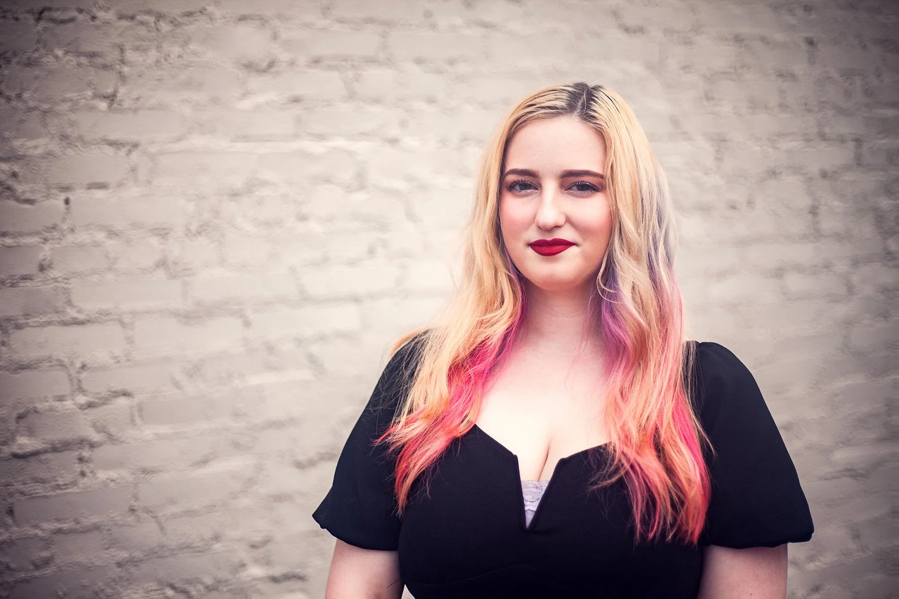 A young woman with blonde hair and pink highlights, wearing a black top, standing against a white brick wall.