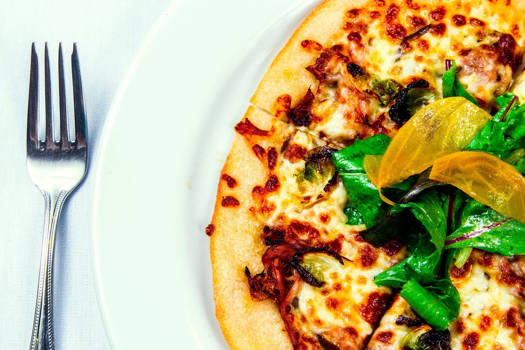 A slice of pizza with melted cheese, pepperoni, and sausage next to a side salad with mixed greens and yellow bell peppers on a white plate, with a fork placed on a white tablecloth.