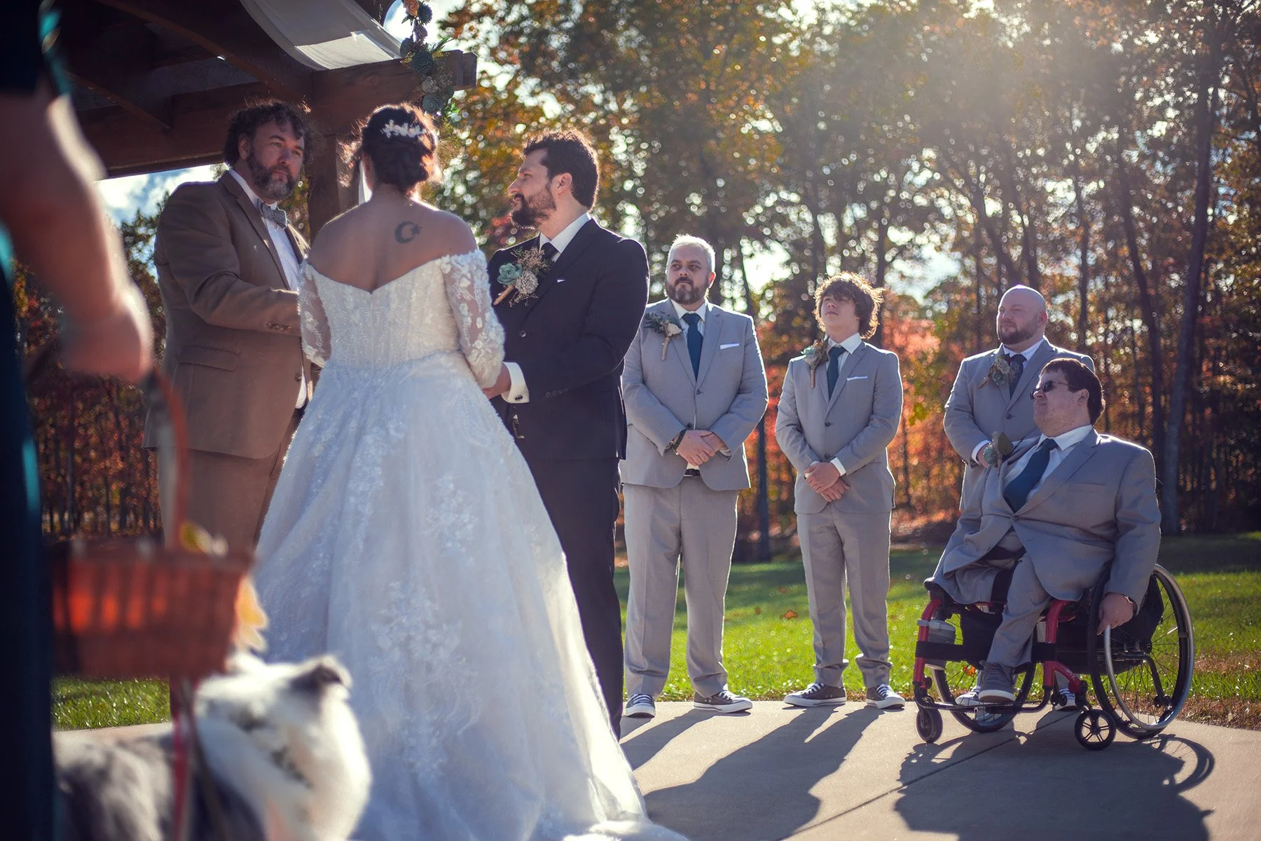 Outdoor wedding ceremony with a bride and groom exchanging vows, surrounded by groomsmen and a man in a wheelchair, with autumn trees in the background.