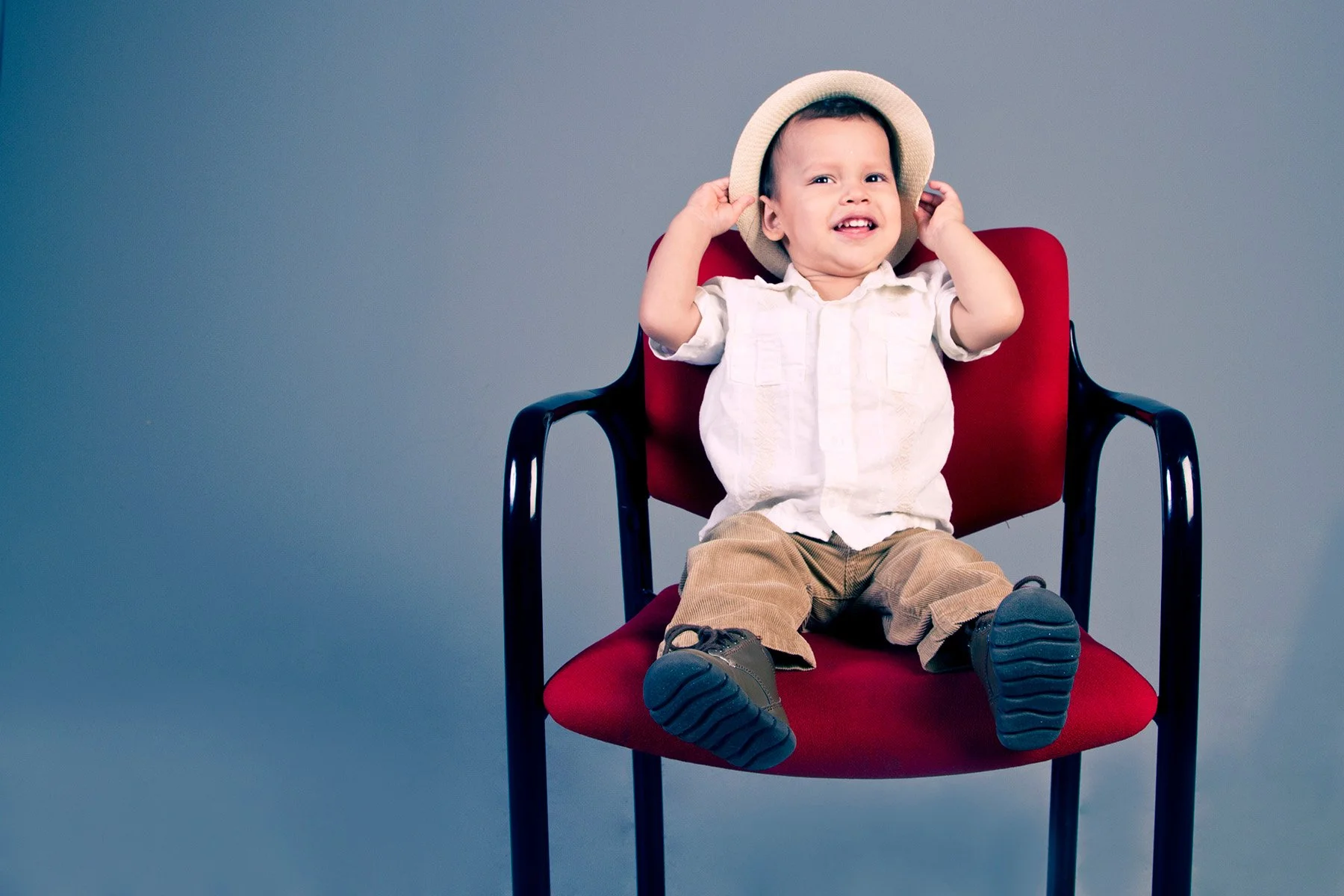 A young boy sitting on a red chair against a grey background, wearing a beige hat, a white shirt, tan pants, and black shoes, smiling and holding the hat.