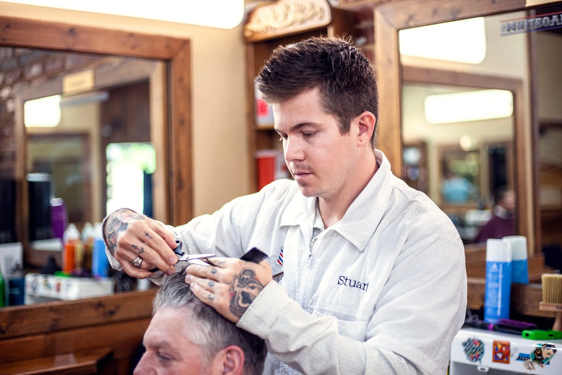 Barber with short dark hair and tattoos on his hands trimming hair of a male client in a barber shop. The barber wears a white shirt with 'Stuart' embroidered on it. The background features wooden framed mirrors and various hair products.