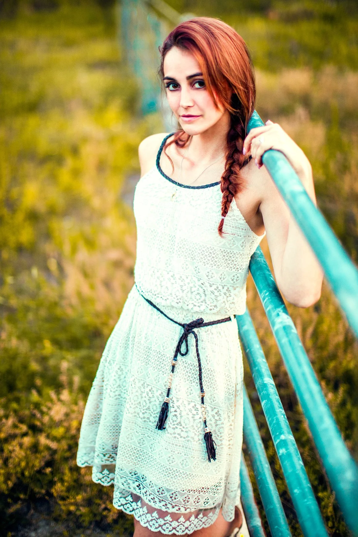 A young woman with red hair braided over her shoulder, wearing a white lace dress with black trim and a black waist tie, standing outdoors and leaning on a blue metal railing.