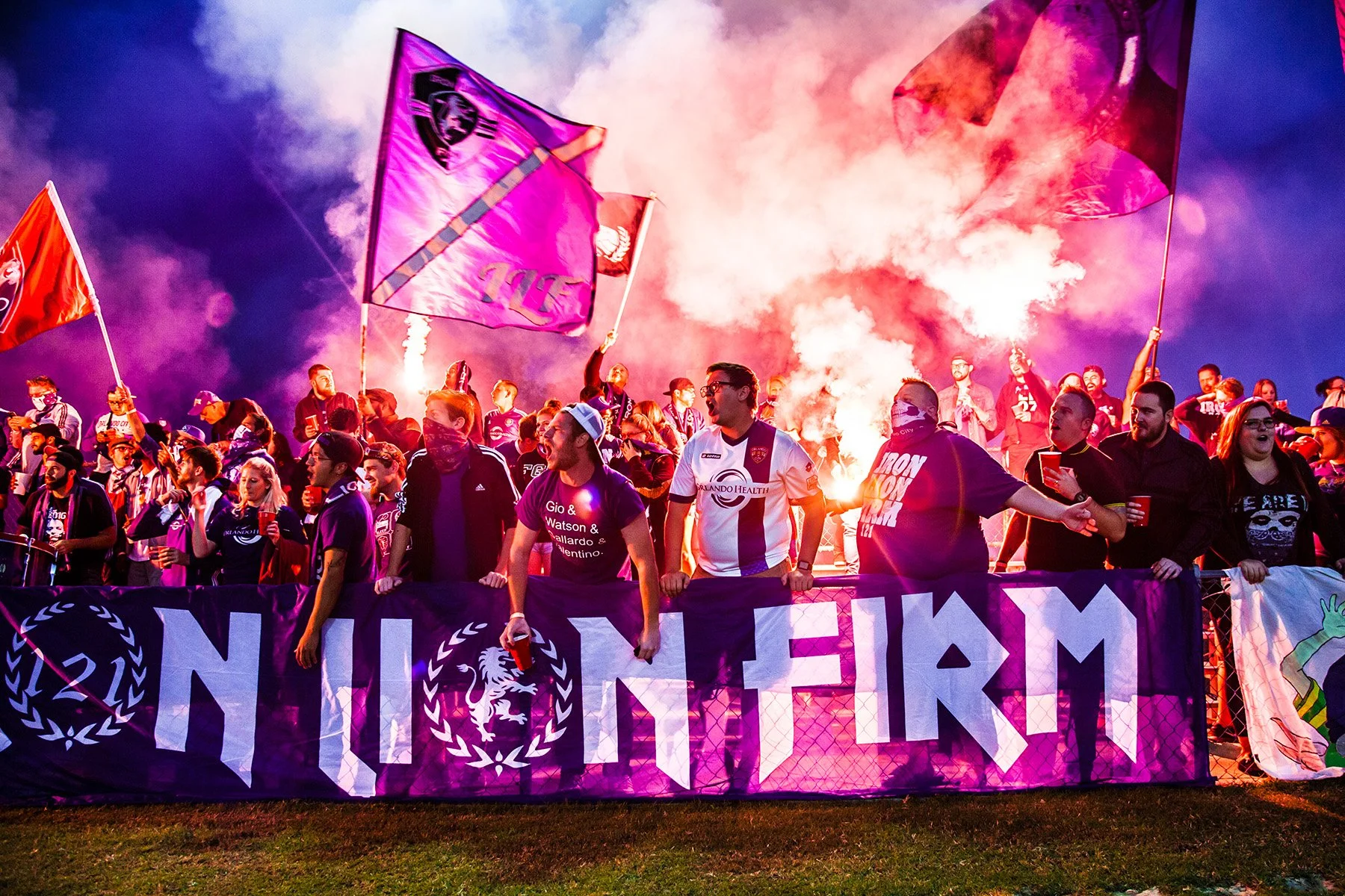 A large group of soccer fans celebrating with flags, smoke, and cheers behind a banner that reads 'Iron Lion Firm,' at twilight or night.