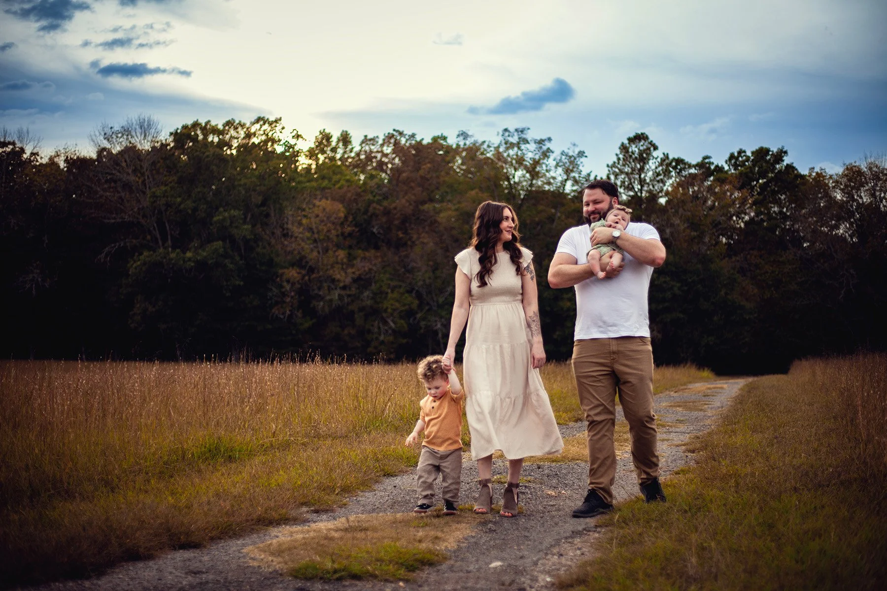 A family of four walking on a dirt path through a field with tall grass and trees in the background during sunset. The father is carrying a baby, the mother is holding the hand of a young boy, and they are all smiling.