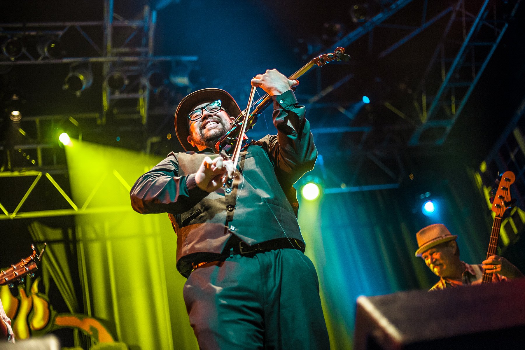 Man wearing glasses and a hat playing violin on stage with colorful stage lights behind him.