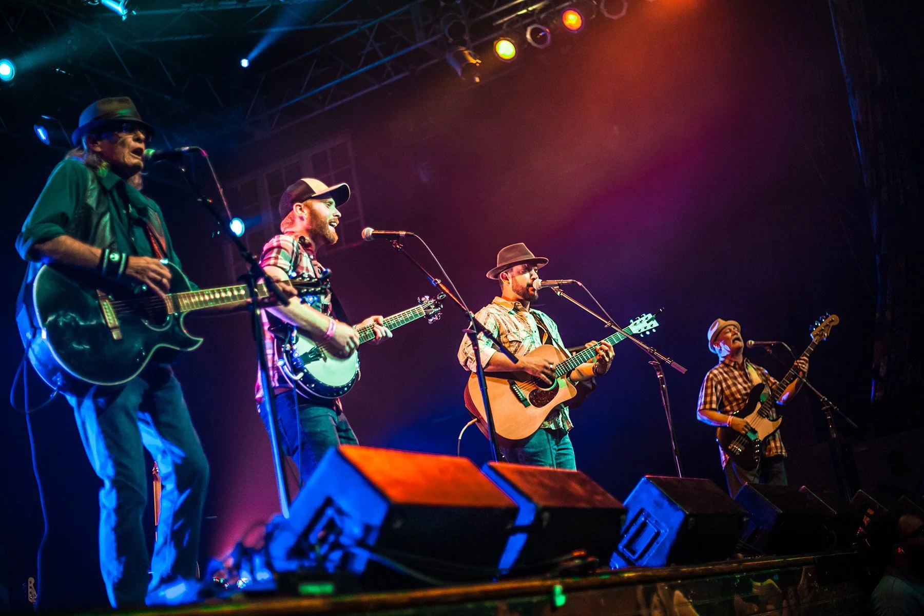 A band of five male musicians performs on stage, playing acoustic and electric guitars, with microphones in front of each. They are dressed casually, some wearing hats, under colorful stage lighting.