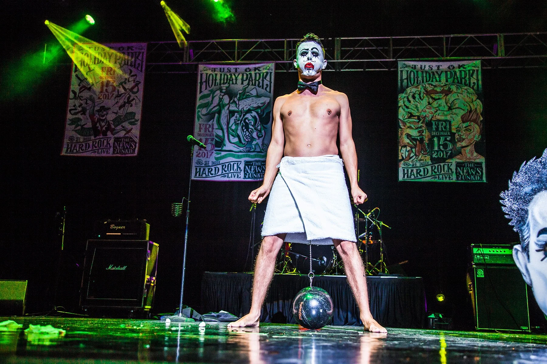 A shirtless man with clown makeup, wearing a bow tie and a towel around his waist, standing on stage with legs apart and a shiny ball under his feet, with flags and musical equipment in the background at a concert or event.