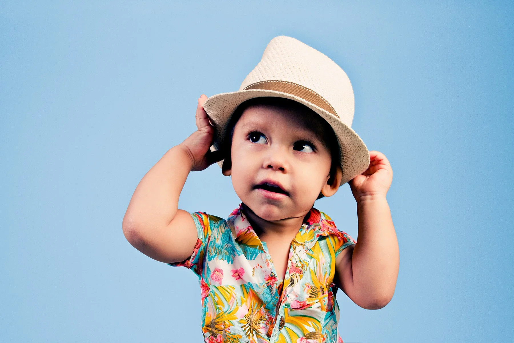 Young child wearing a colorful tropical shirt and a beige straw hat, adjusting the hat with both hands against a solid light blue background.