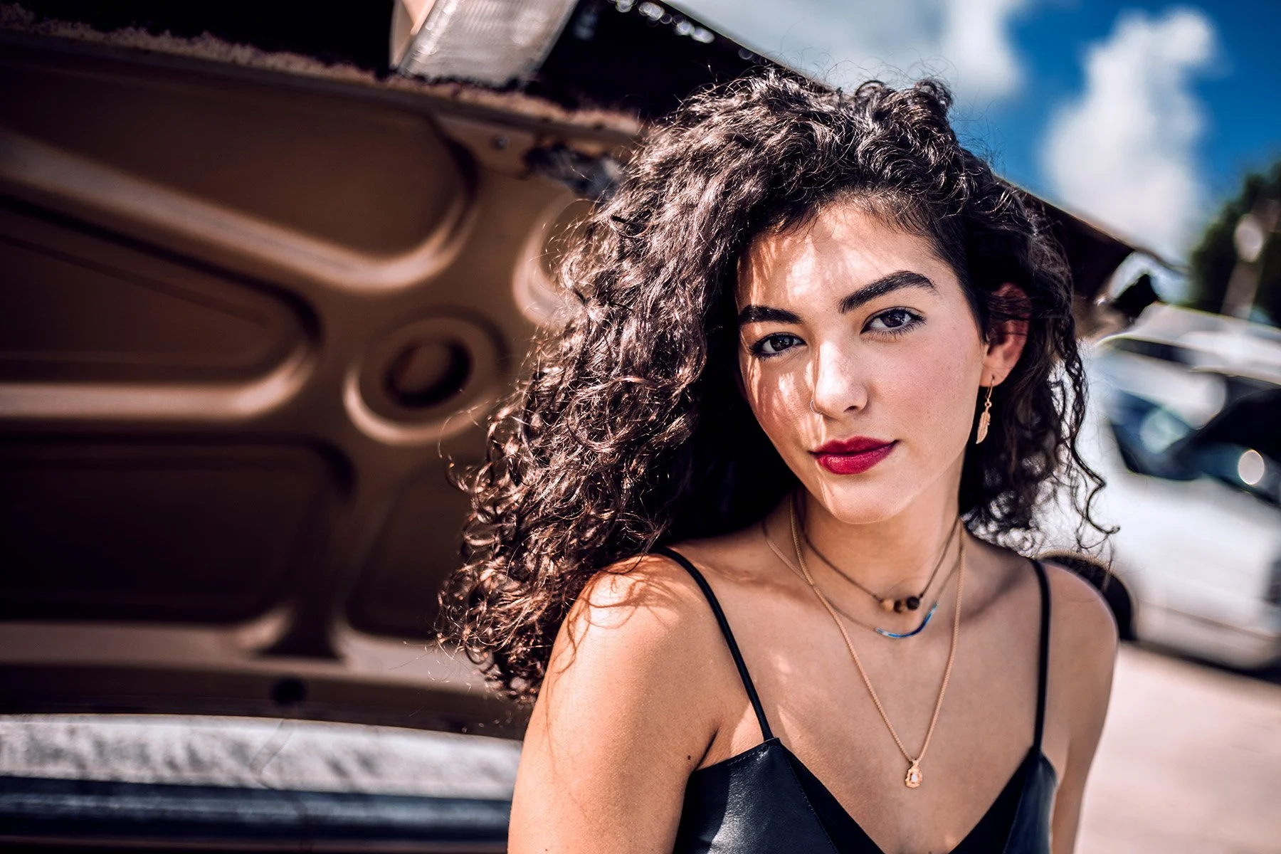 Young woman with curly dark hair, red lipstick, and jewelry, leaning against the open trunk of a car outdoors on a sunny day.