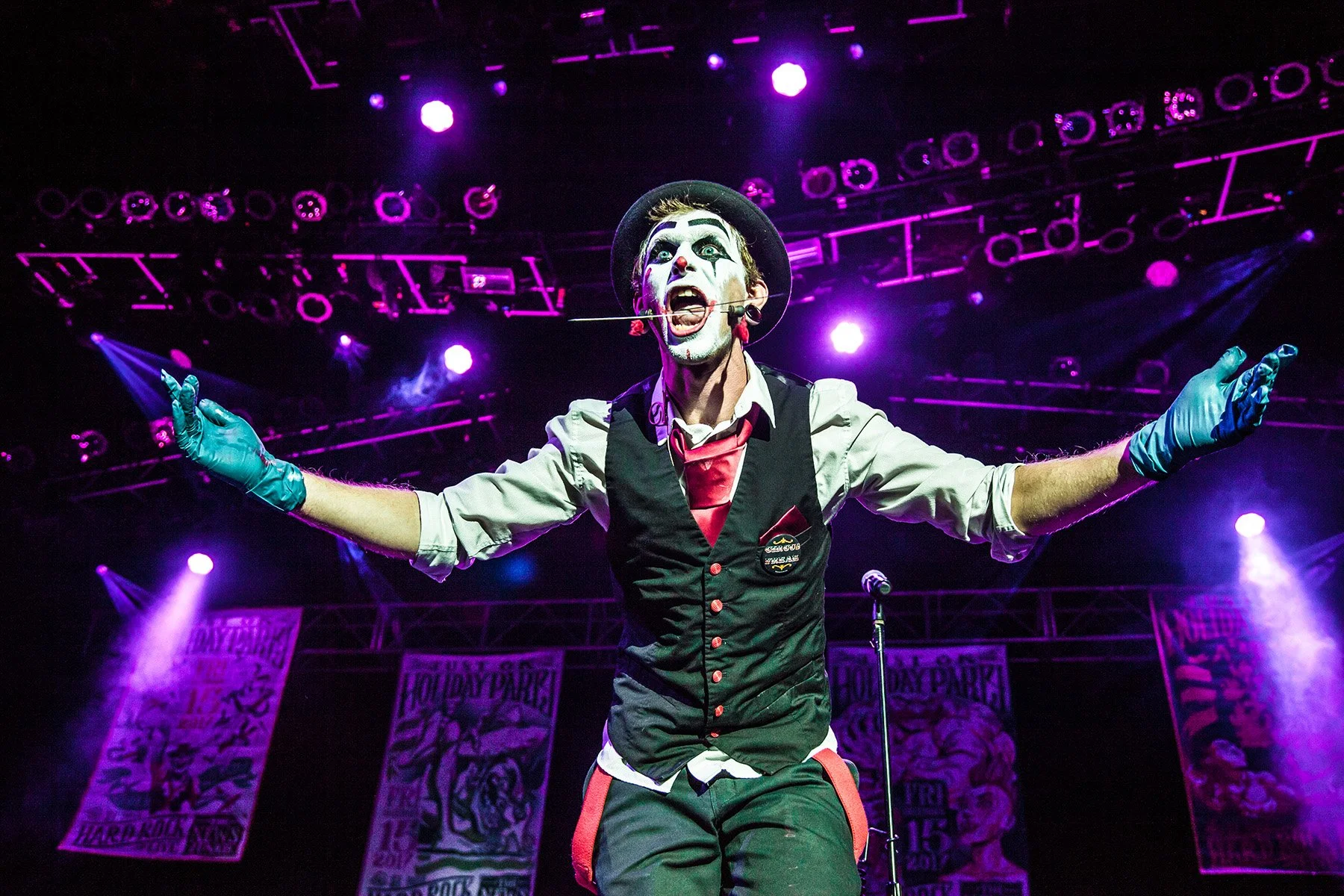 Performer on stage with arms outstretched, wearing a black hat, makeup, a black vest over a white shirt, and blue gloves, under purple stage lighting.