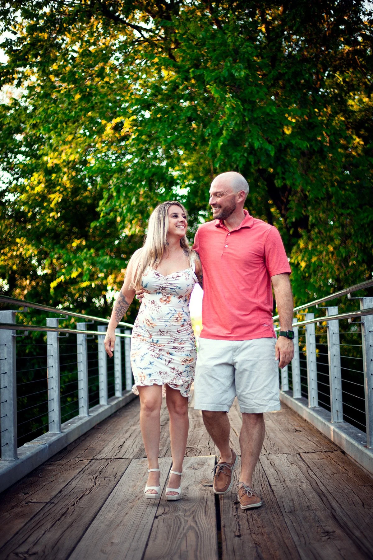 A smiling couple walking together on a wooden bridge surrounded by green trees.