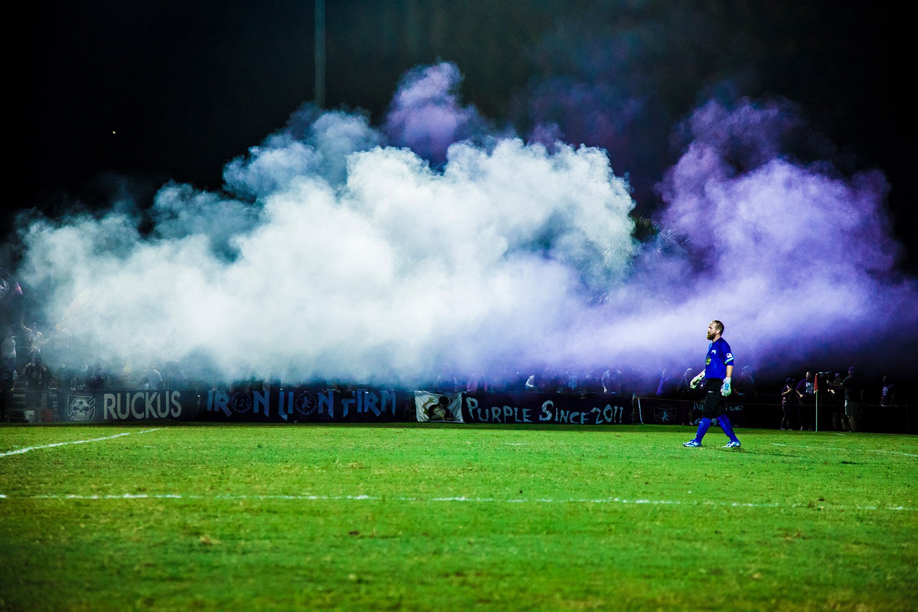 A soccer stadium at night with purple and white smoke filling the air behind the goalpost. A soccer goalie in a blue uniform walks on the field. Banners with text are displayed along the sideline.