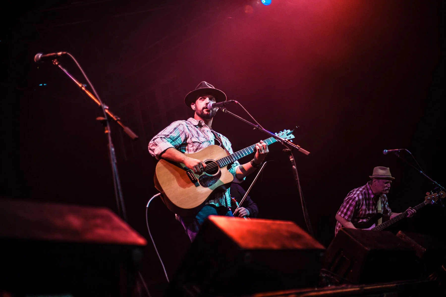 A male musician wearing a hat and plaid shirt playing an acoustic guitar and singing into a microphone on stage during a concert, with stage lights creating a red glow.