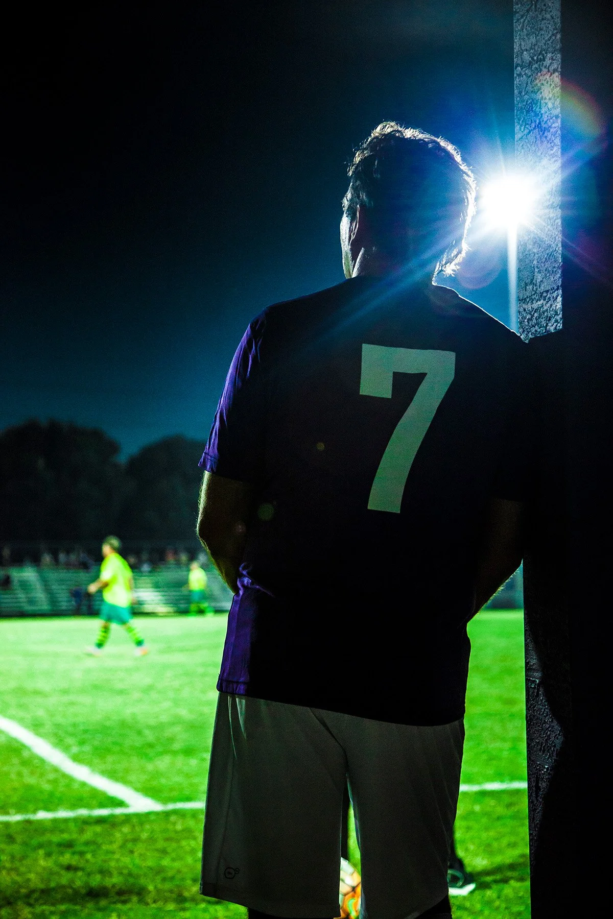 A soccer player wearing a jersey with the number 7 stands near the sideline during a night game, illuminated by stadium lights, with other players visible in the background on the field.