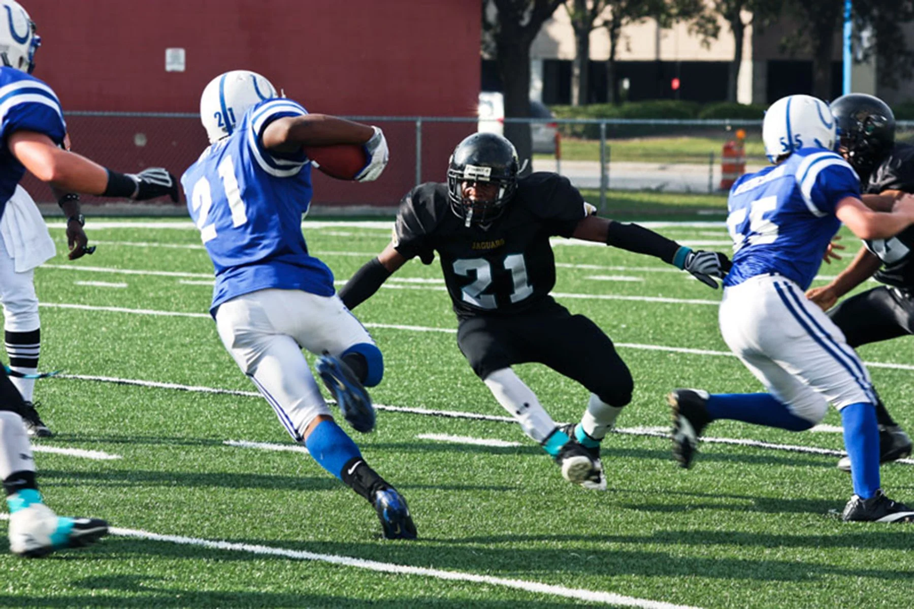 A youth football game with players in blue and black uniforms on an artificial turf field. One player in blue is holding the football and running, while a black-uniformed player attempts to block him. Other players and trees are visible in the backgr