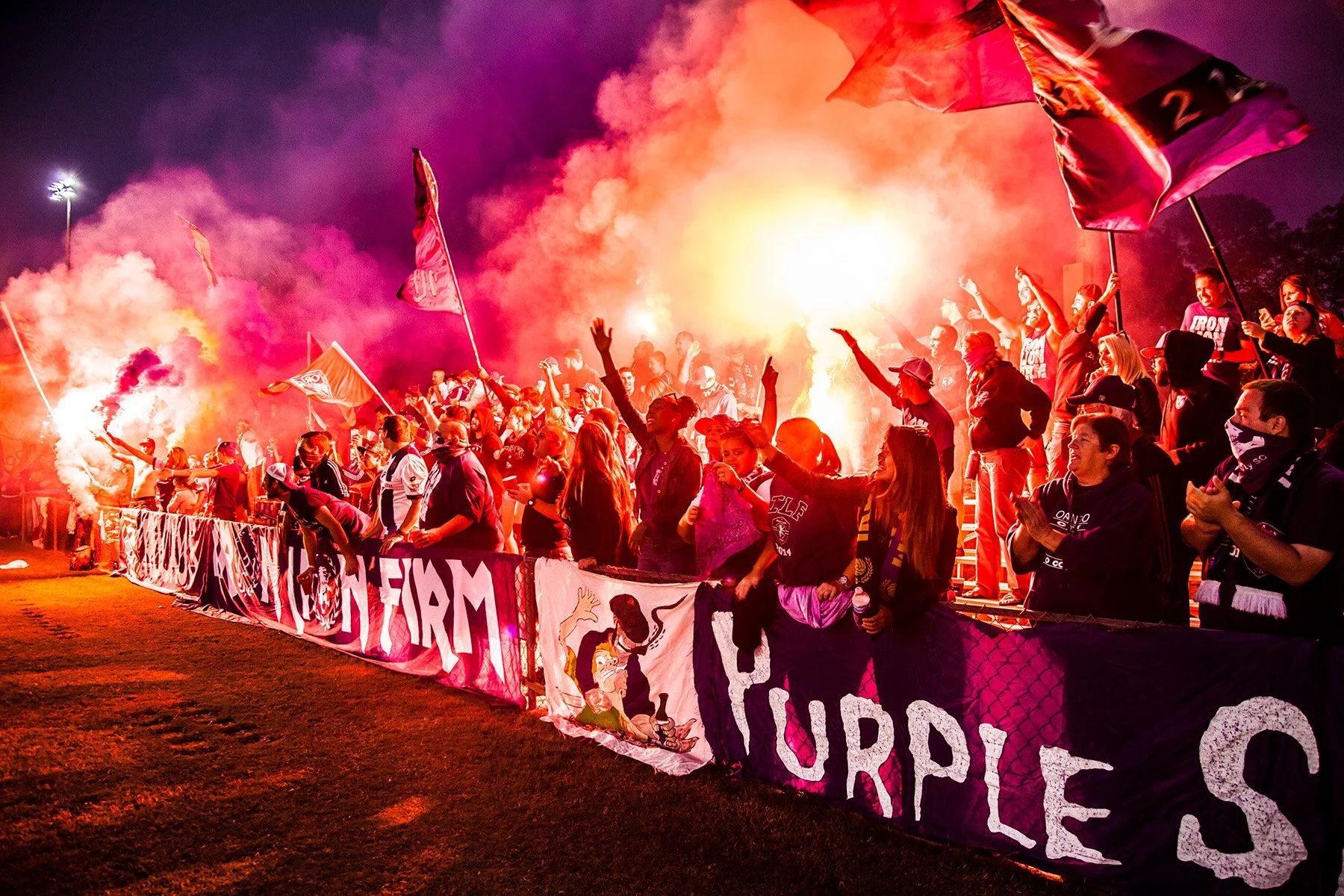 A group of sports fans cheering at a stadium during night time, with some holding flags and flares creating smoke and bright light.