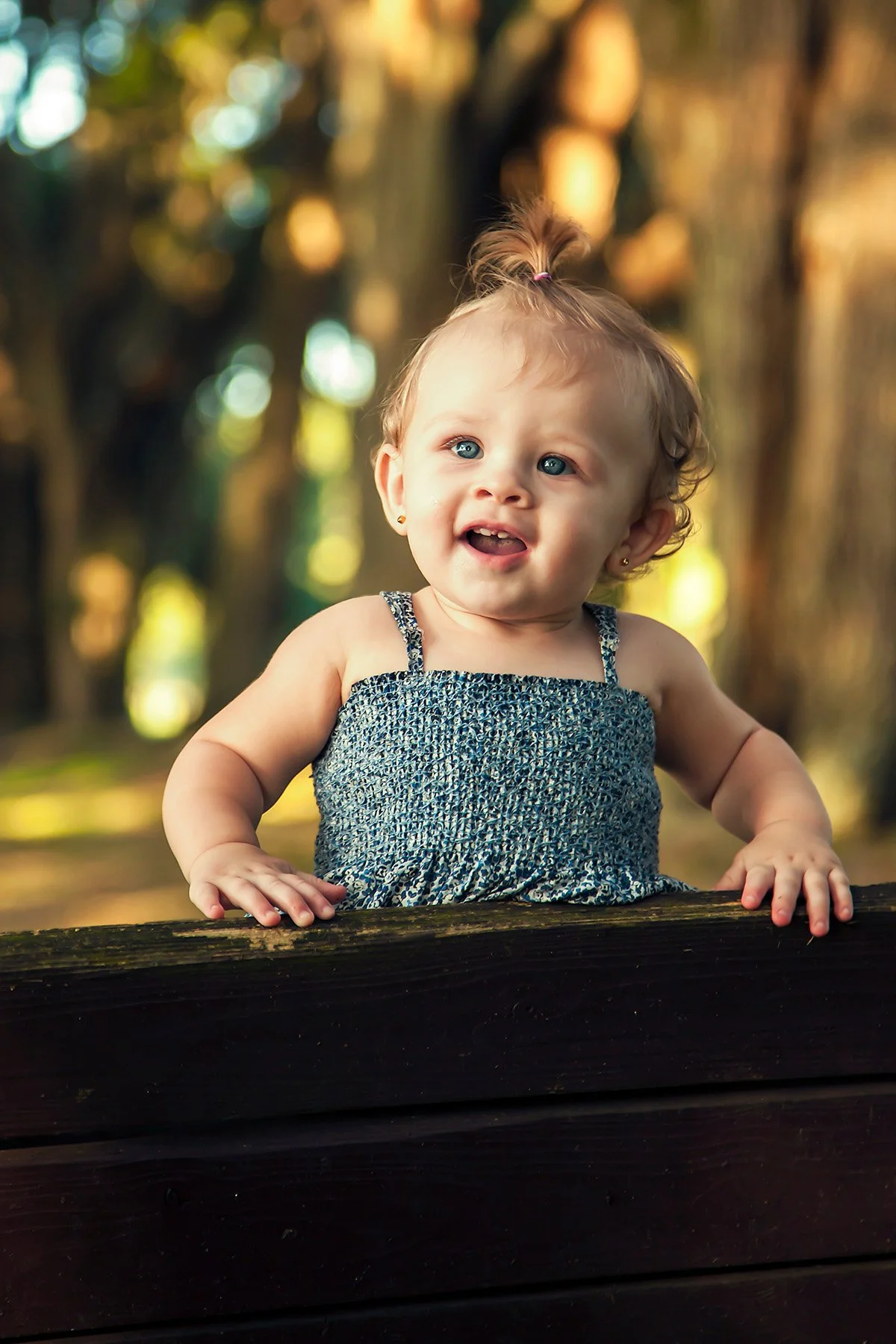 A young girl with big blue eyes and light curly hair in a small ponytail, wearing a blue patterned dress, is leaning on a wooden rail outdoors with a blurred background of trees and dappled sunlight.