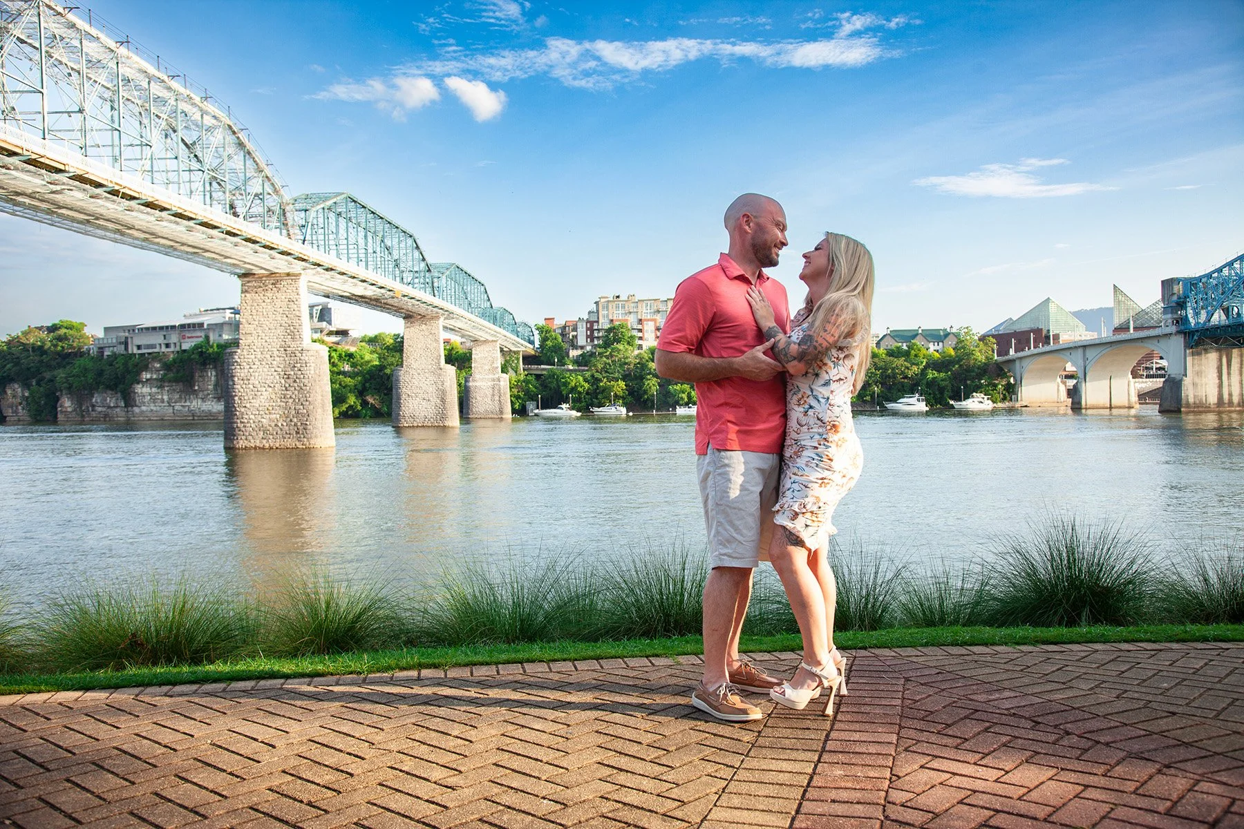 A couple standing by the river, smiling and looking into each other's eyes. The man is wearing a pink shirt and beige shorts, while the woman is wearing a floral dress and high heels. There are bridges and boats on the river in the background, with g