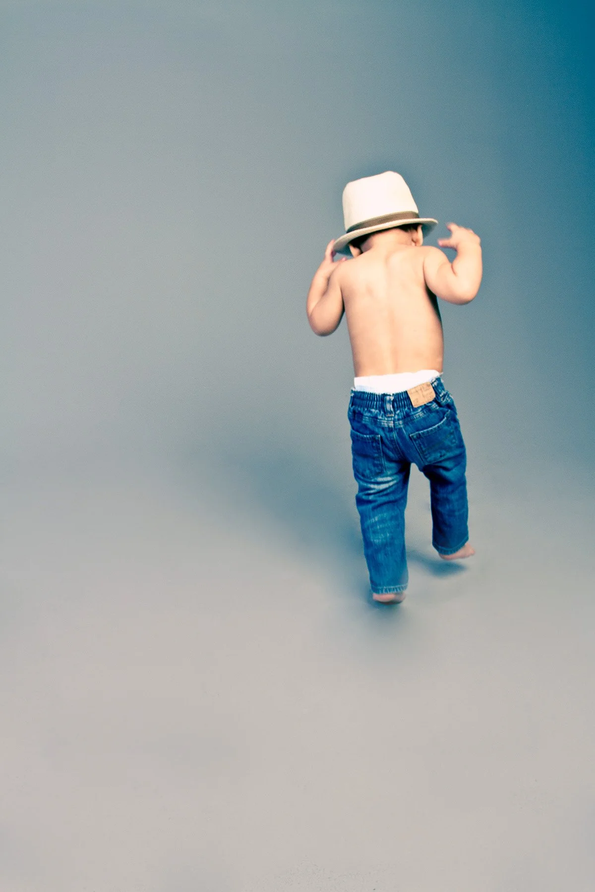 A shirtless child wearing jeans and a white fedora hat, walking barefoot against a plain background.