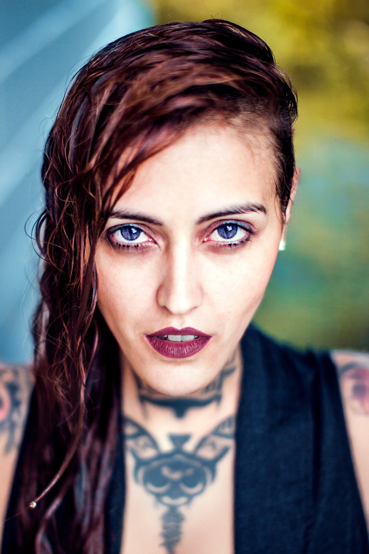 A woman with blue eyes and dark red hair styled to one side, wearing dark lipstick, with visible tattoos on her chest and arms, looking directly at the camera.