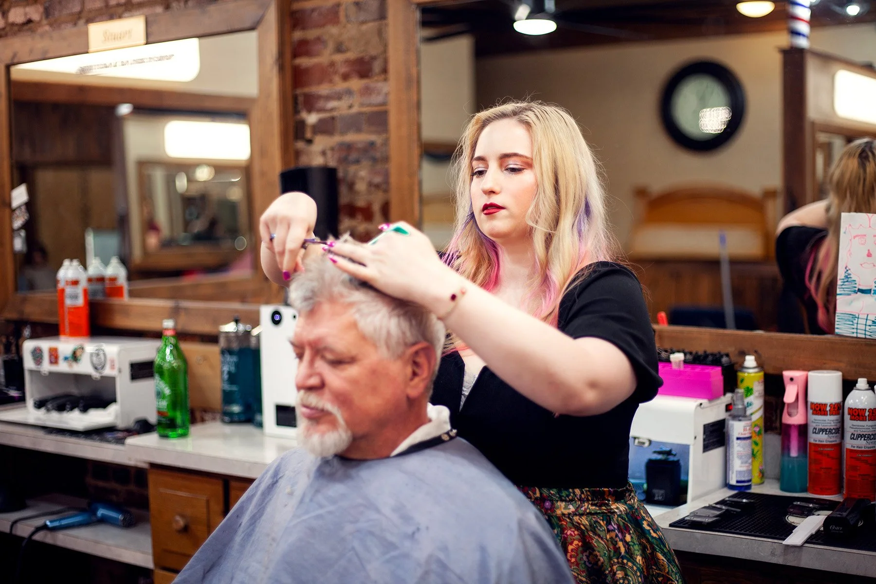 A young woman with blonde hair and pink highlights is cutting a man's hair in a barbershop. The man has gray hair and a beard and is seated. The barbershop has wooden accents, mirrors, and various hair care products on the counter.