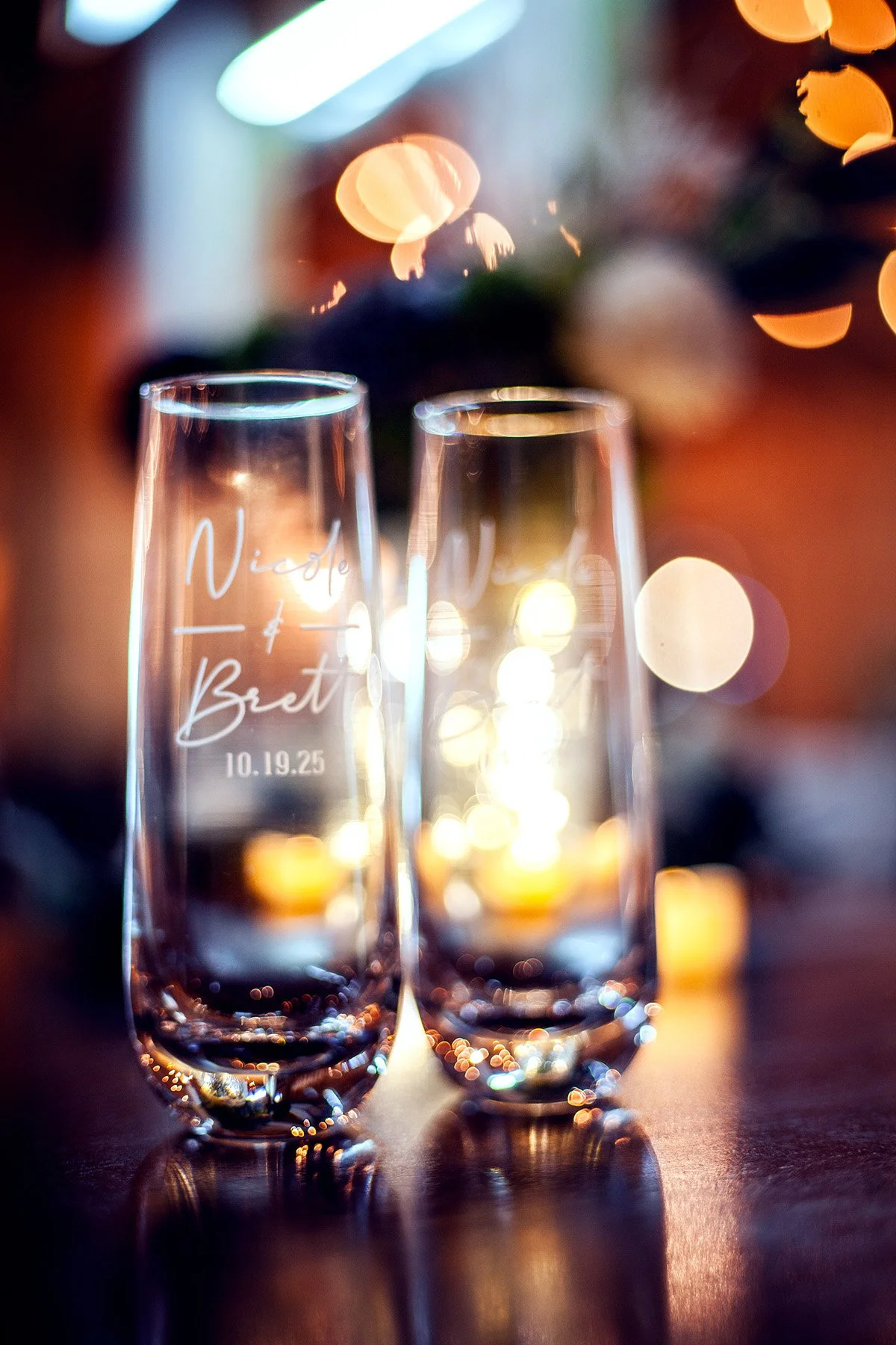 Two champagne flutes with engraved names 'Nicole & Brett' and date '10.19.25', reflecting ambient lights and bokeh background.