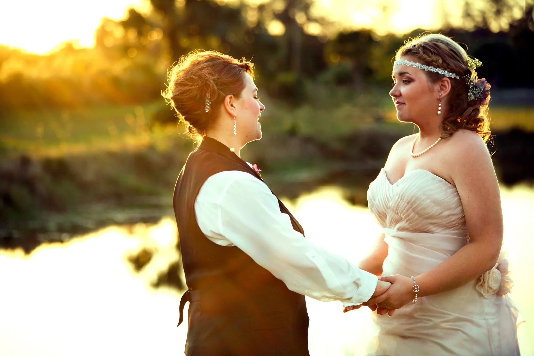 A same-sex couple holding hands and facing each other during their wedding ceremony at sunset outdoors, with a water body and trees in the background.