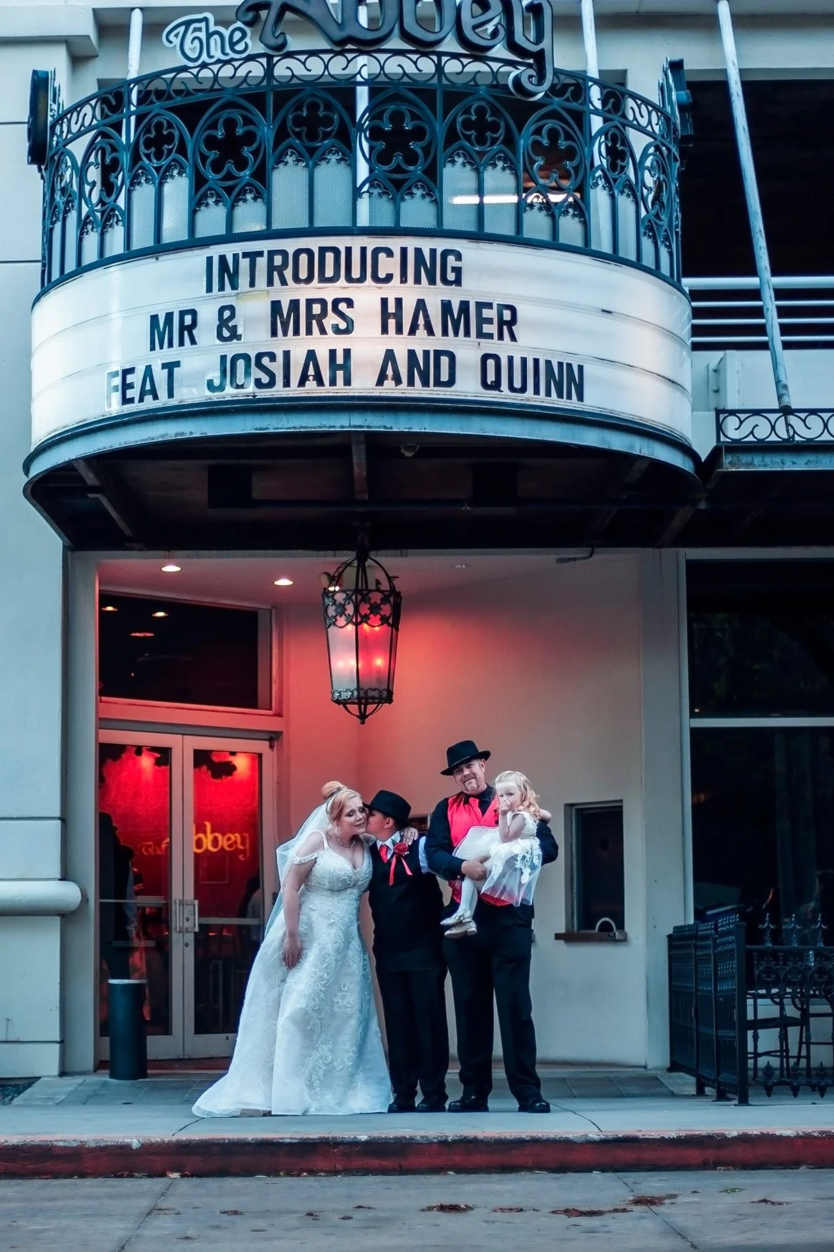 A wedding scene outside a theater with a sign introducing Mr. and Mrs. Hamer and their children, Josiah and Quinn. There is a bride in a white wedding dress and veil, a groom in a black suit, and two children in formal attire. The setting includes a 