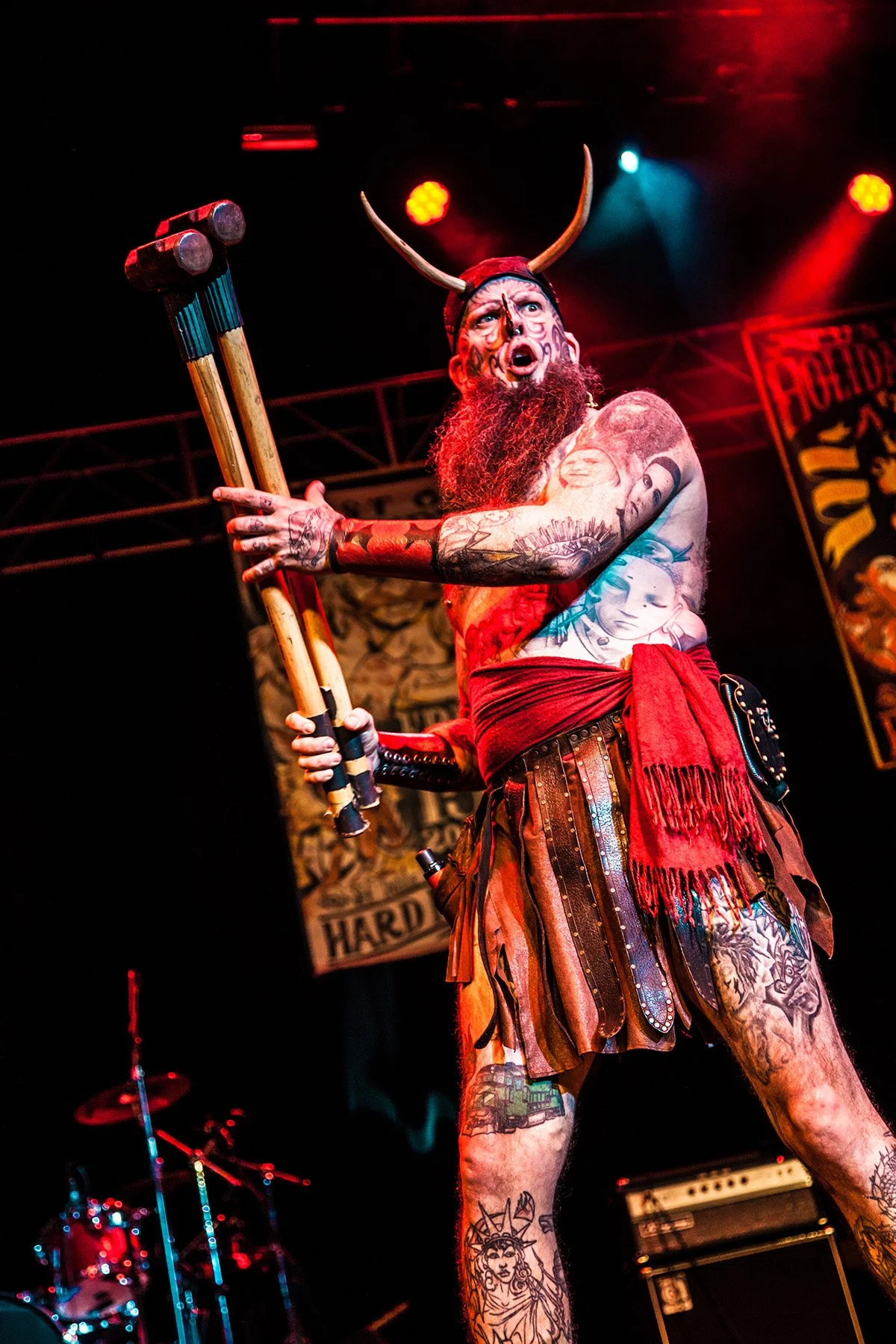 Person dressed as a Viking warrior with horned helmet, tattoos, and traditional clothing, holding a sledgehammer on a stage with red and blue lighting.