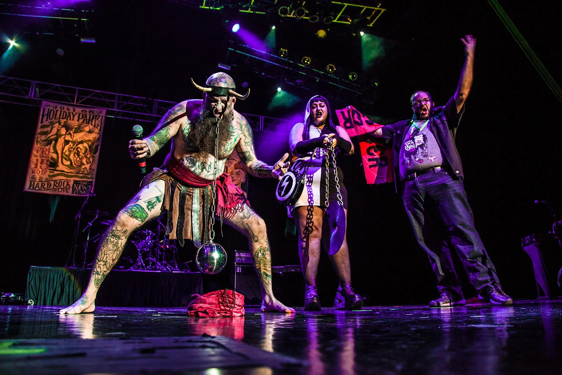 Three performers on stage during a concert with colorful purple and green lighting. The first performer on the left is dressed in tribal costume with tattoos, a Viking helmet with horns, and holding a microphone. The middle performer is a woman dress