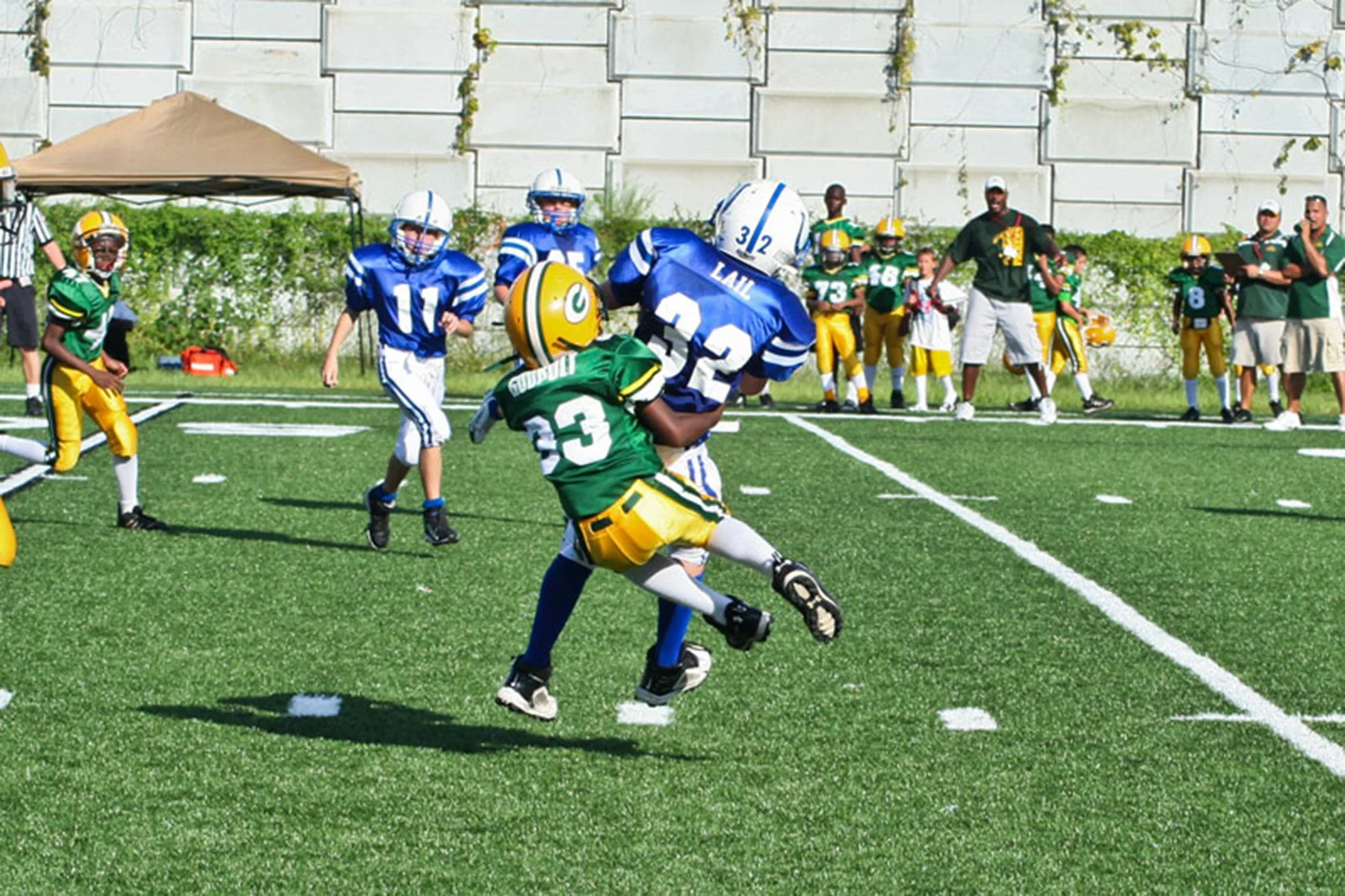 Young football players in green and yellow and blue and white uniforms playing a game on a green football field, with spectators and coaches watching from the sidelines.