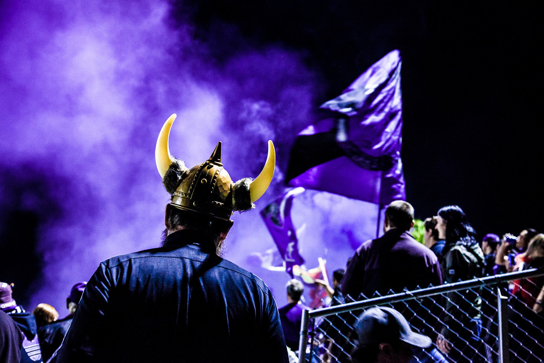 A crowd of people at a sports event, with one person wearing a Viking helmet with horns and a dark jacket, watching a male athlete in purple apparel climbing a rope or ladder during a nighttime event with purple lighting and smoke effects.