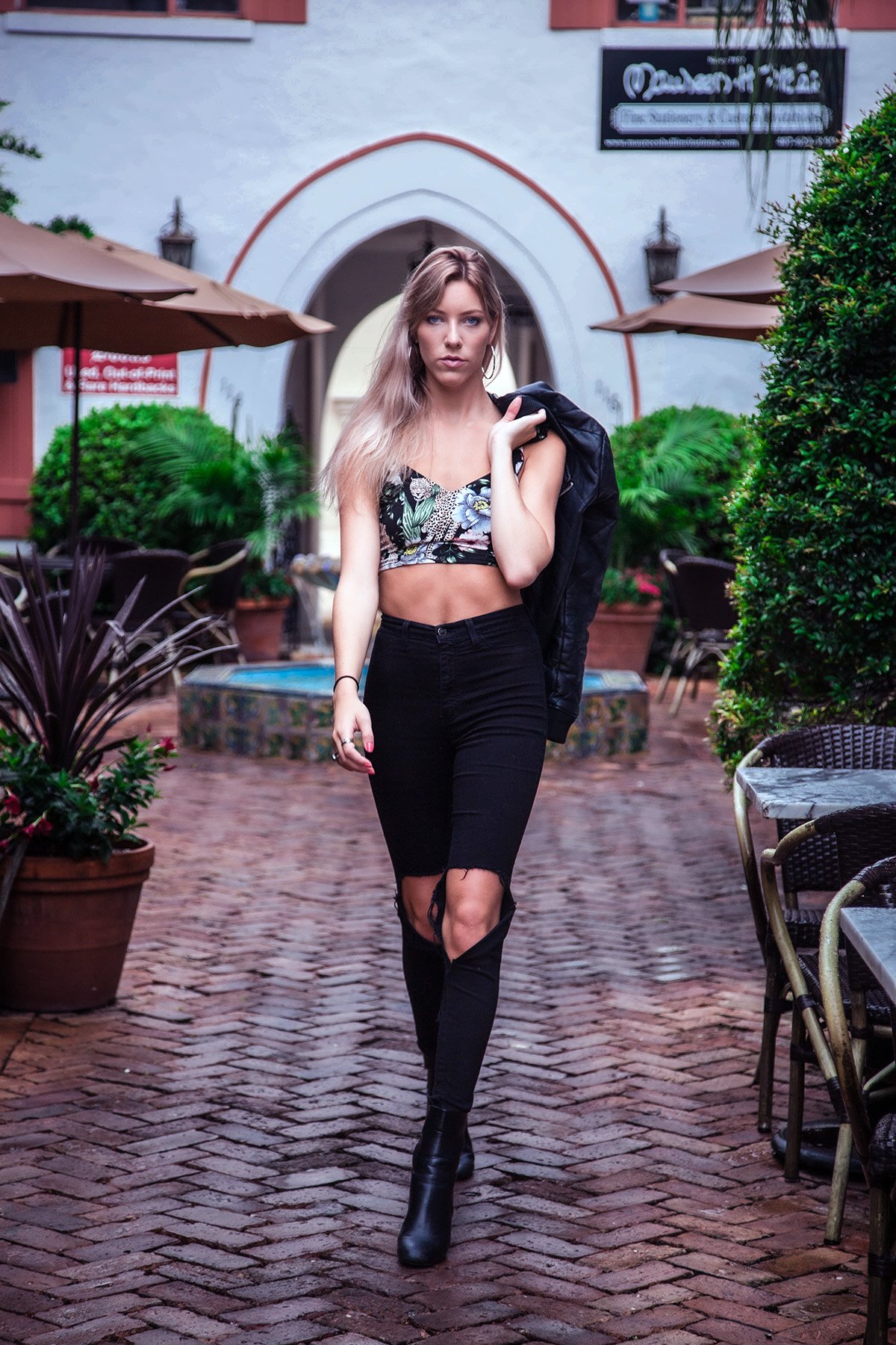 A young woman walking on a brick patio with potted plants and outdoor seating, wearing a floral crop top, ripped black jeans, and boots, carrying a black jacket over her shoulder.