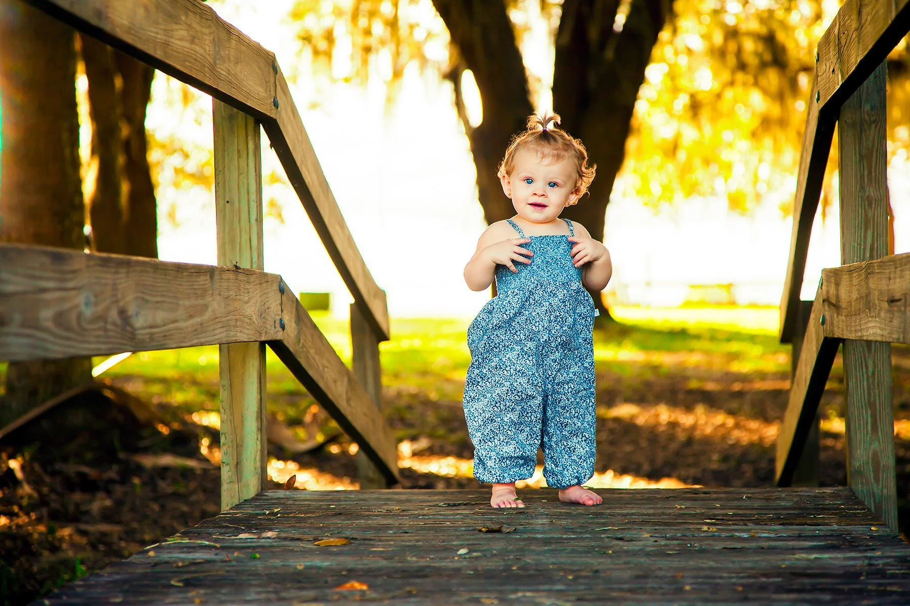A young girl with curly hair and blue eyes standing barefoot on a wooden bridge outdoors, wearing a blue floral jumpsuit with a background of trees with yellow-orange autumn leaves.