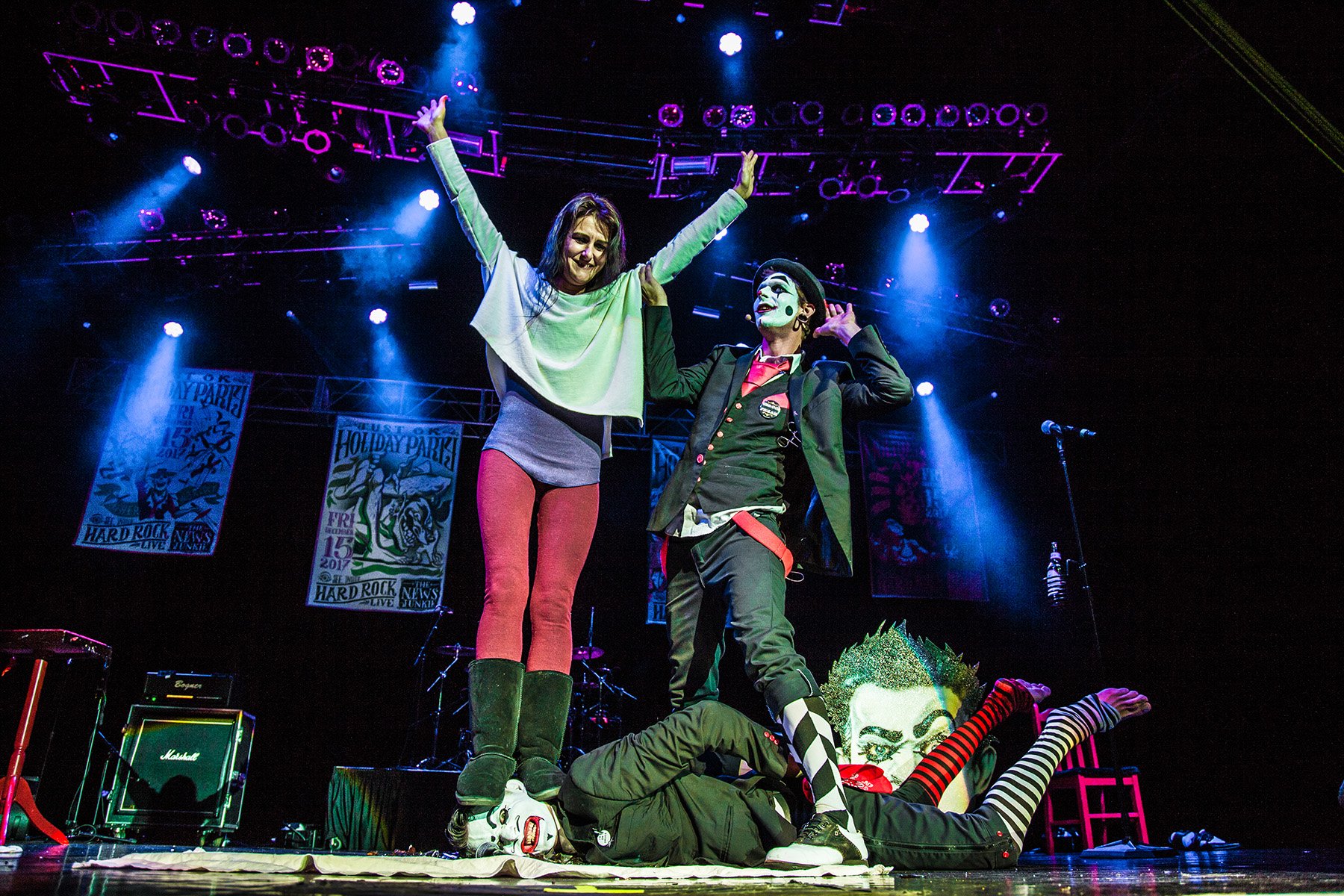 A theatrical stage performance featuring two clowns dressed in colorful costumes and makeup, with a woman in a casual sweater, red pants, and green boots, raising her arms with a joyful expression. The stage is lit with purple and blue lights, with p