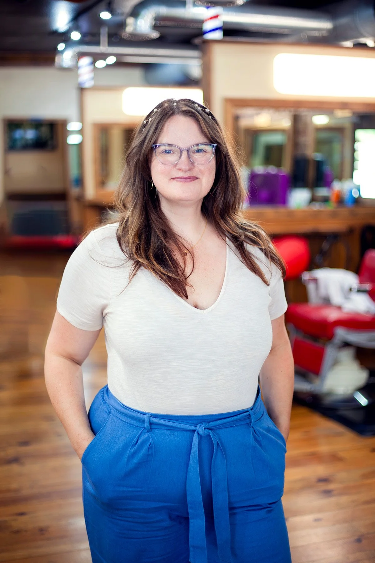 A woman with long brown hair, wearing glasses, a white V-neck T-shirt, and blue pants with a drawstring, standing in a barbershop or salon, smiling with her hands in her pockets.
