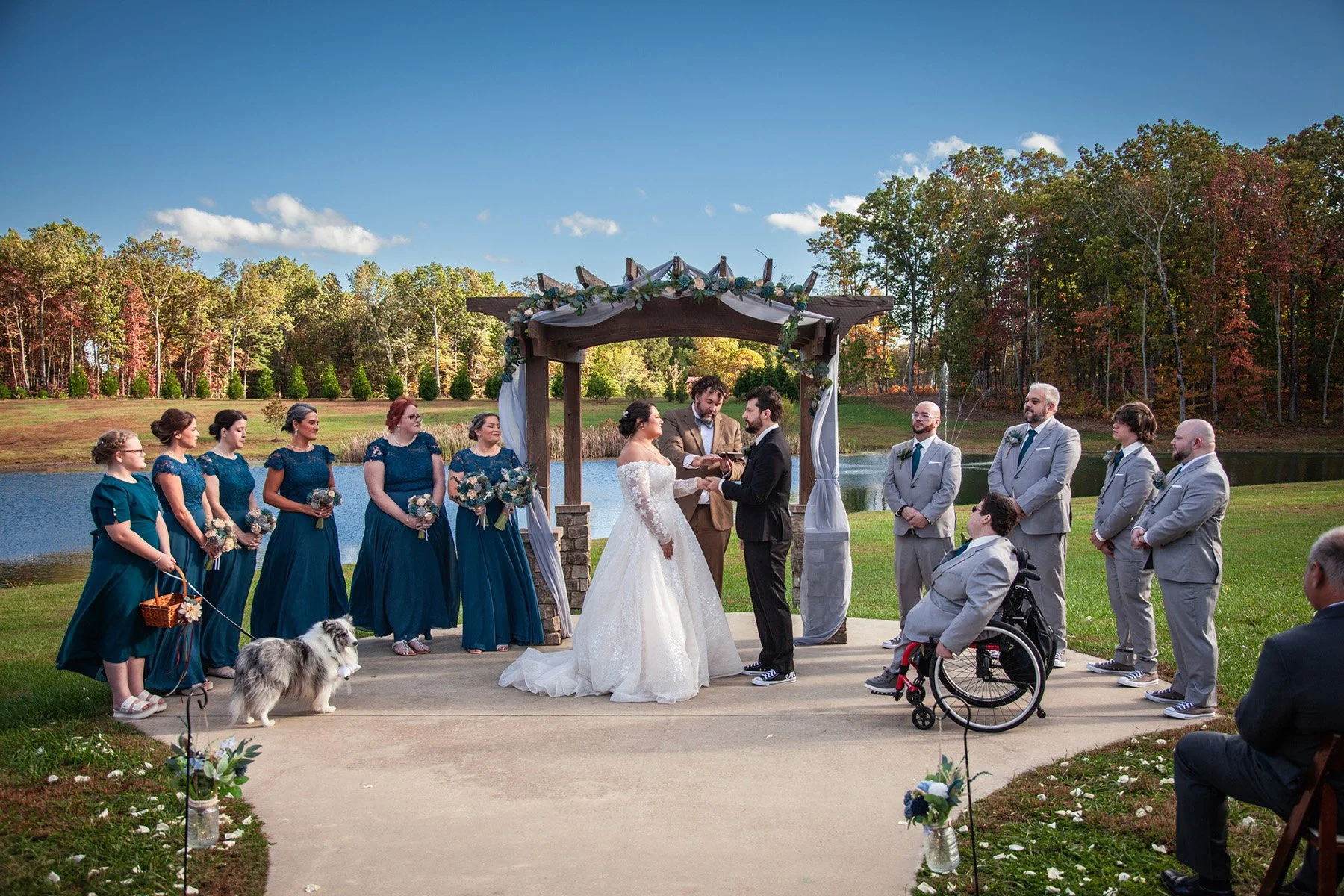Outdoor wedding ceremony with a bride and groom exchanging vows under a decorated wooden arch, surrounded by wedding party and guests, near a lake with trees in the background on a sunny day.