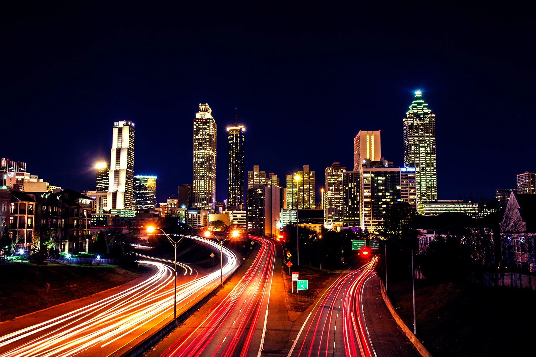 Night view of a city skyline with illuminated skyscrapers and light trails from moving vehicles on the highway in the foreground.