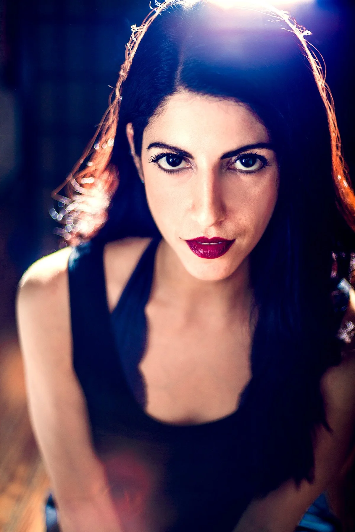 Close-up of a woman with dark hair, blue eyes, and dark red lipstick looking directly at the camera, with sunlight shining behind her head.
