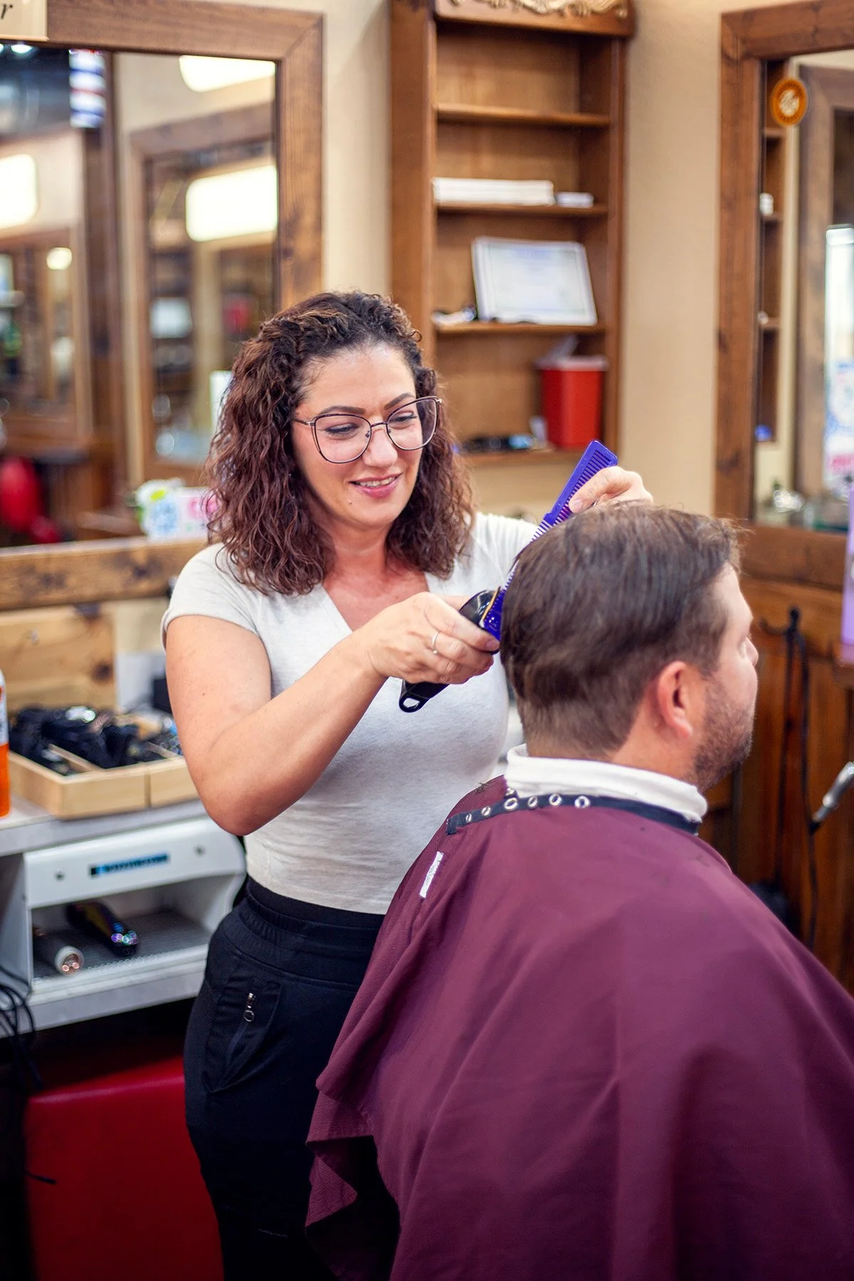 A woman is cutting a man's hair in a barber shop, surrounded by wooden mirrors and shelves.